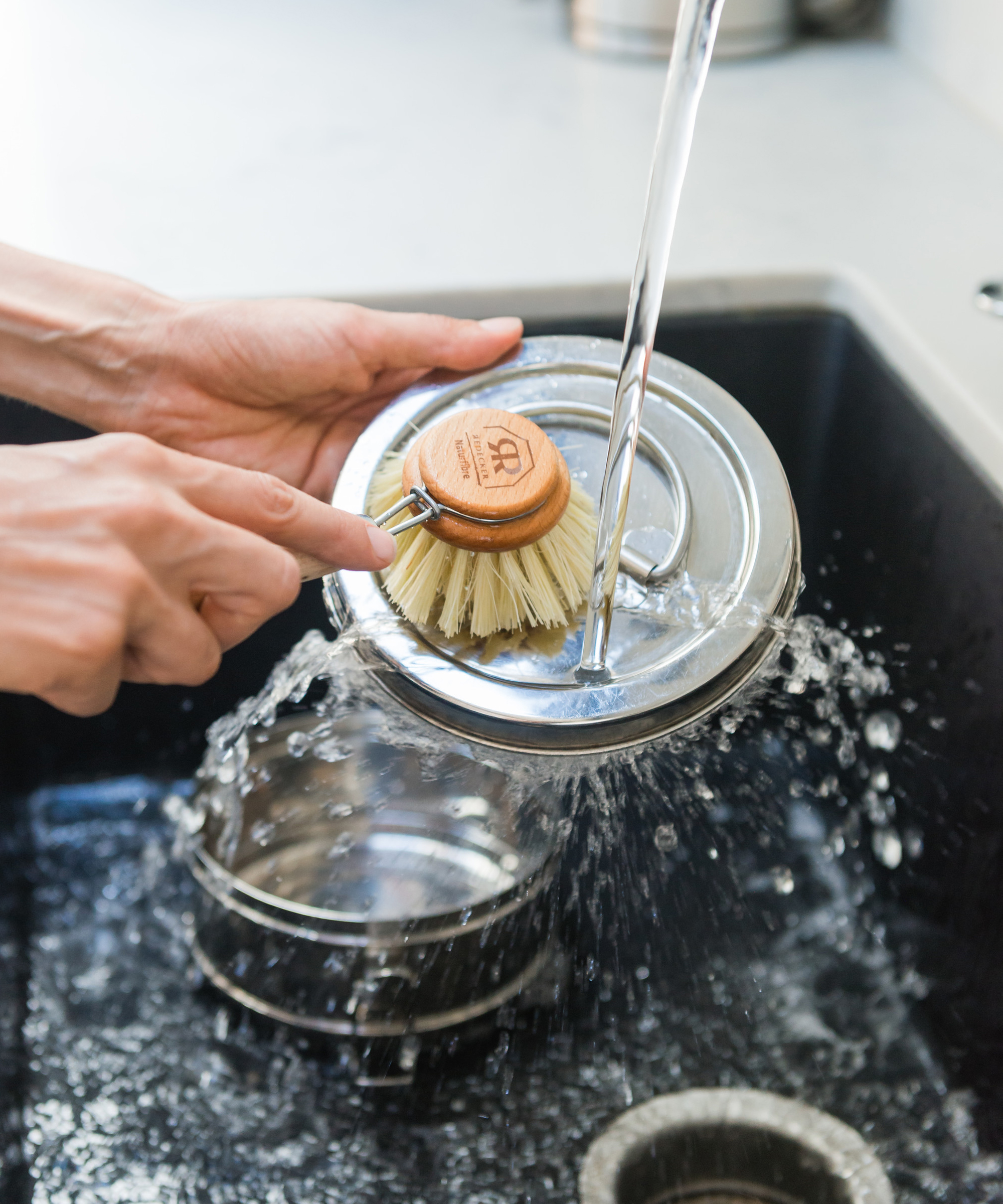 A wild minimalist effortlessly cleans a metal pot lid under running water, using a wooden dish brush. Other metal items rest in the sink below, highlighting a simple, uncluttered lifestyle.