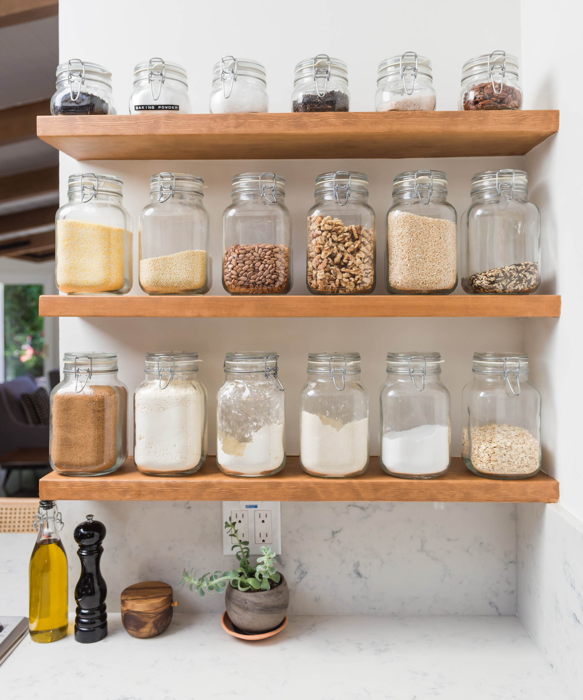 Three wooden shelves in this wild minimalist kitchen hold glass jars filled with grains, nuts, and sugar. Below, a marble counter displays olive oil, a pepper grinder, and a small potted plant against a crisp white wall.
