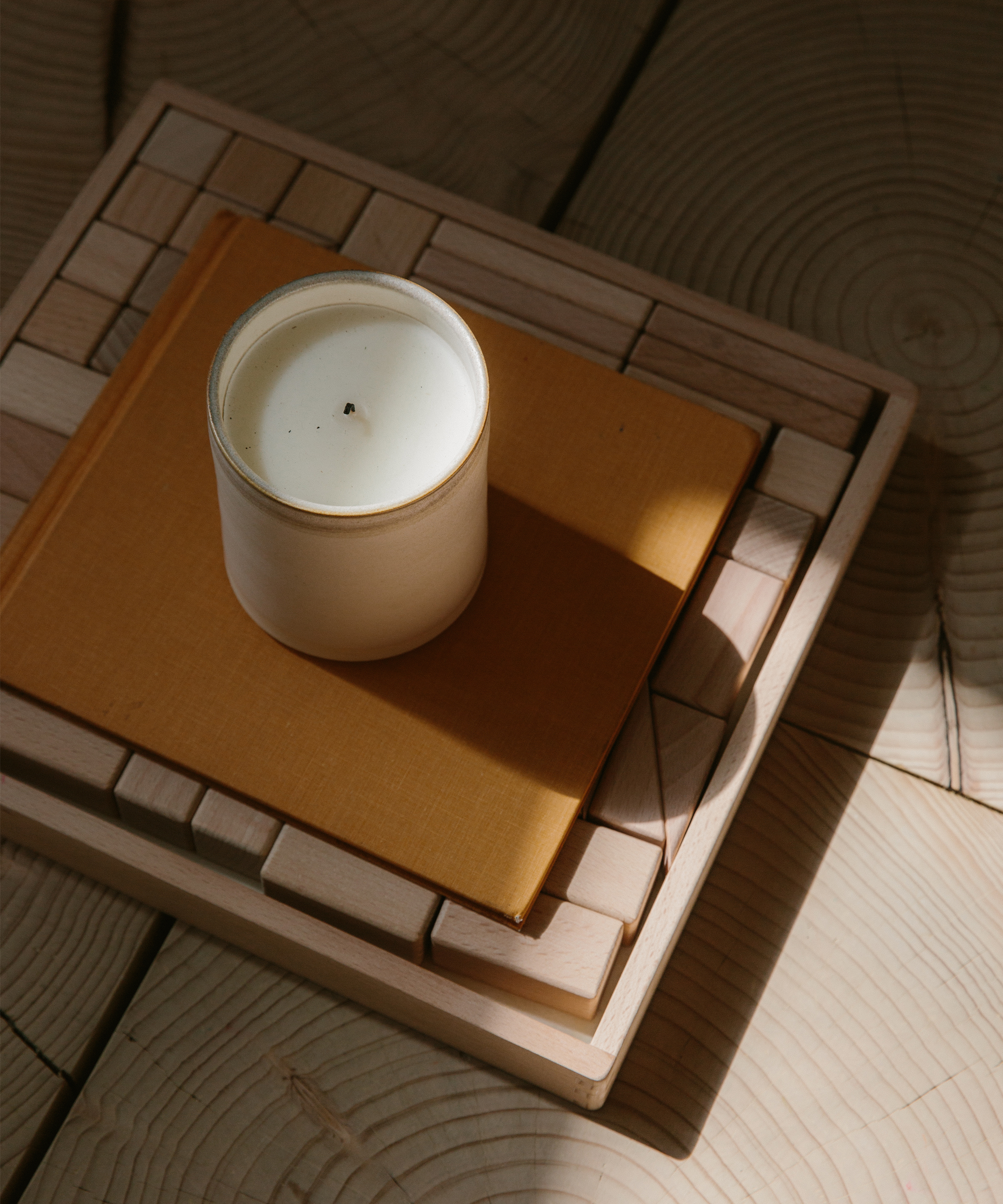A white candle in a glass jar sits on a closed, mustard-colored book, both placed on a decorative wooden tray atop a wooden surface with visible circular grain patterns—perfect for relaxing with an ara katz seed probiotics ritual in soft sunlight.