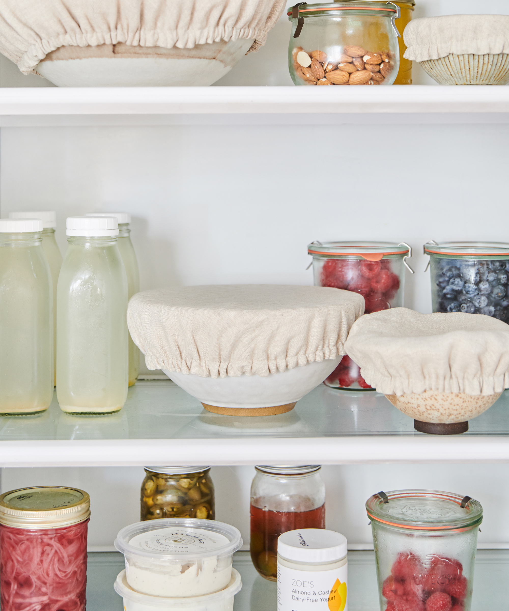 Open refrigerator shelves with jars of preserved foods, glass bottles of light-colored liquid, bowls covered with beige reusable fabric covers, and containers of berries, nuts, and spreads—just like in a Pamela Salzman produce and pantry guide.