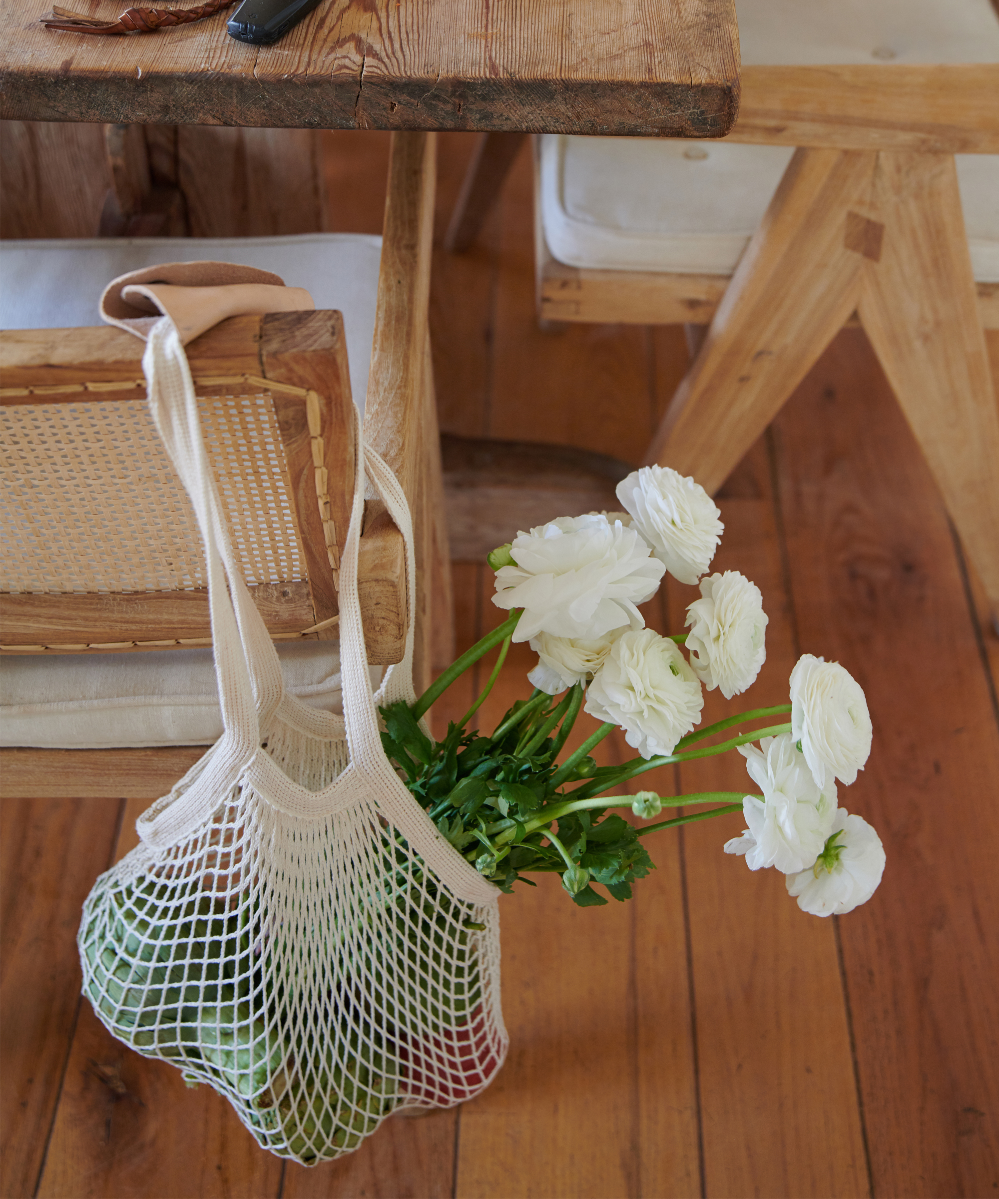 A reusable mesh bag, inspired by the Pamela Salzman produce and pantry guide, brims with green produce and white flowers as it hangs on a wooden chair beside a rustic table atop a wooden floor.