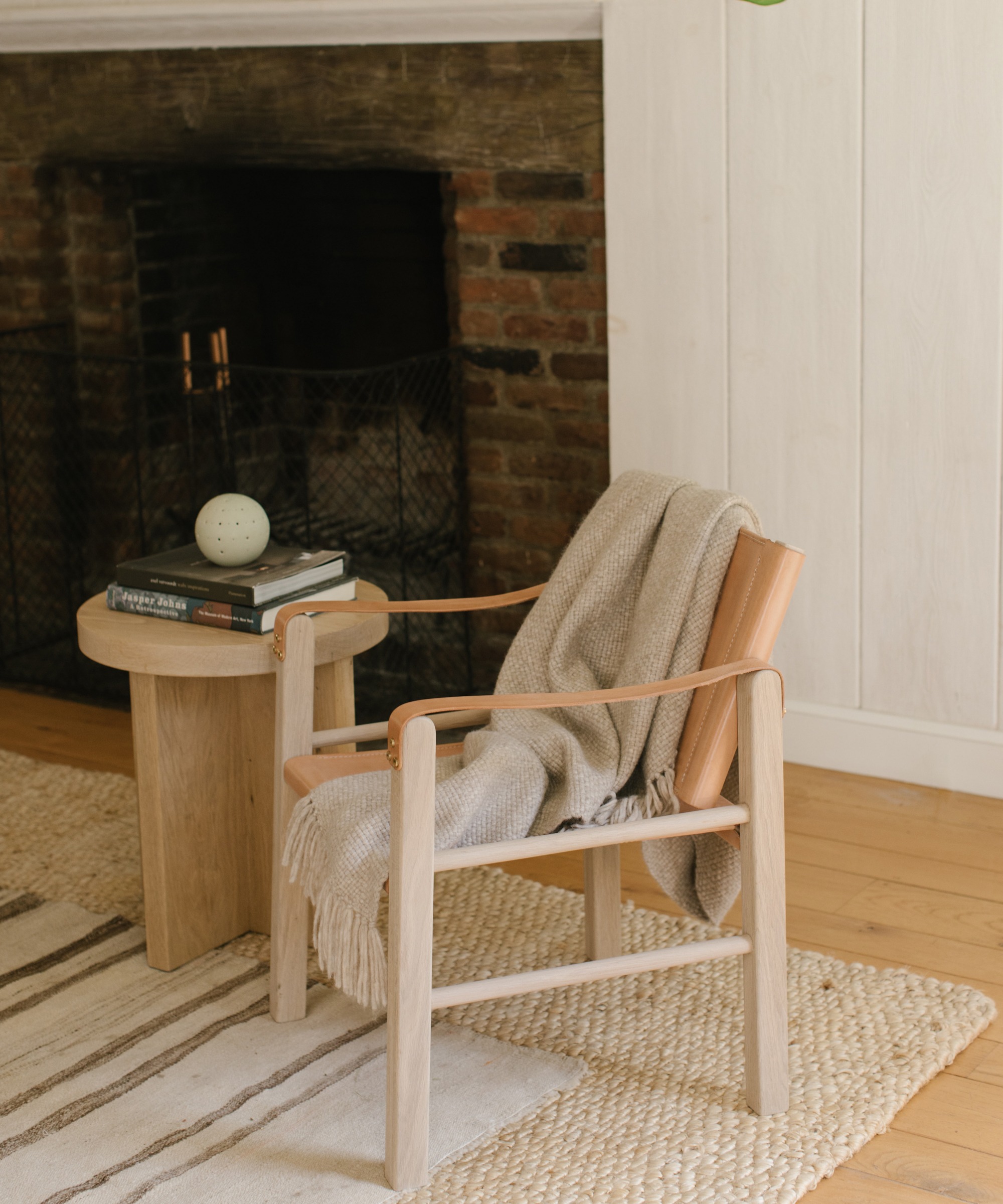 Room interior with a brown chair, tan throw blanket, wood side table, brick fireplace, woven rug, and white walls.