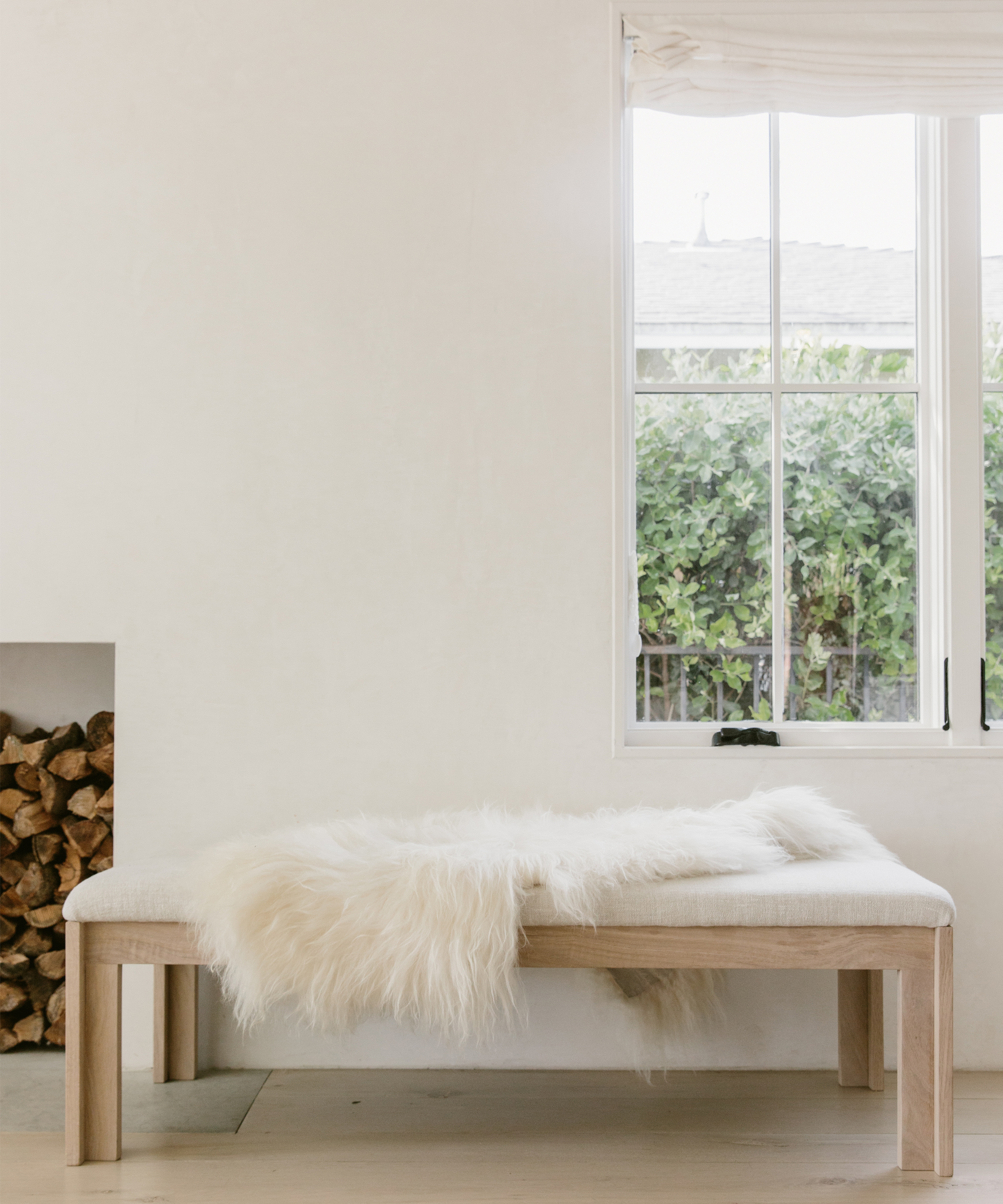Wooden bench with white faux fur throw blanket in front of a window and white walls.