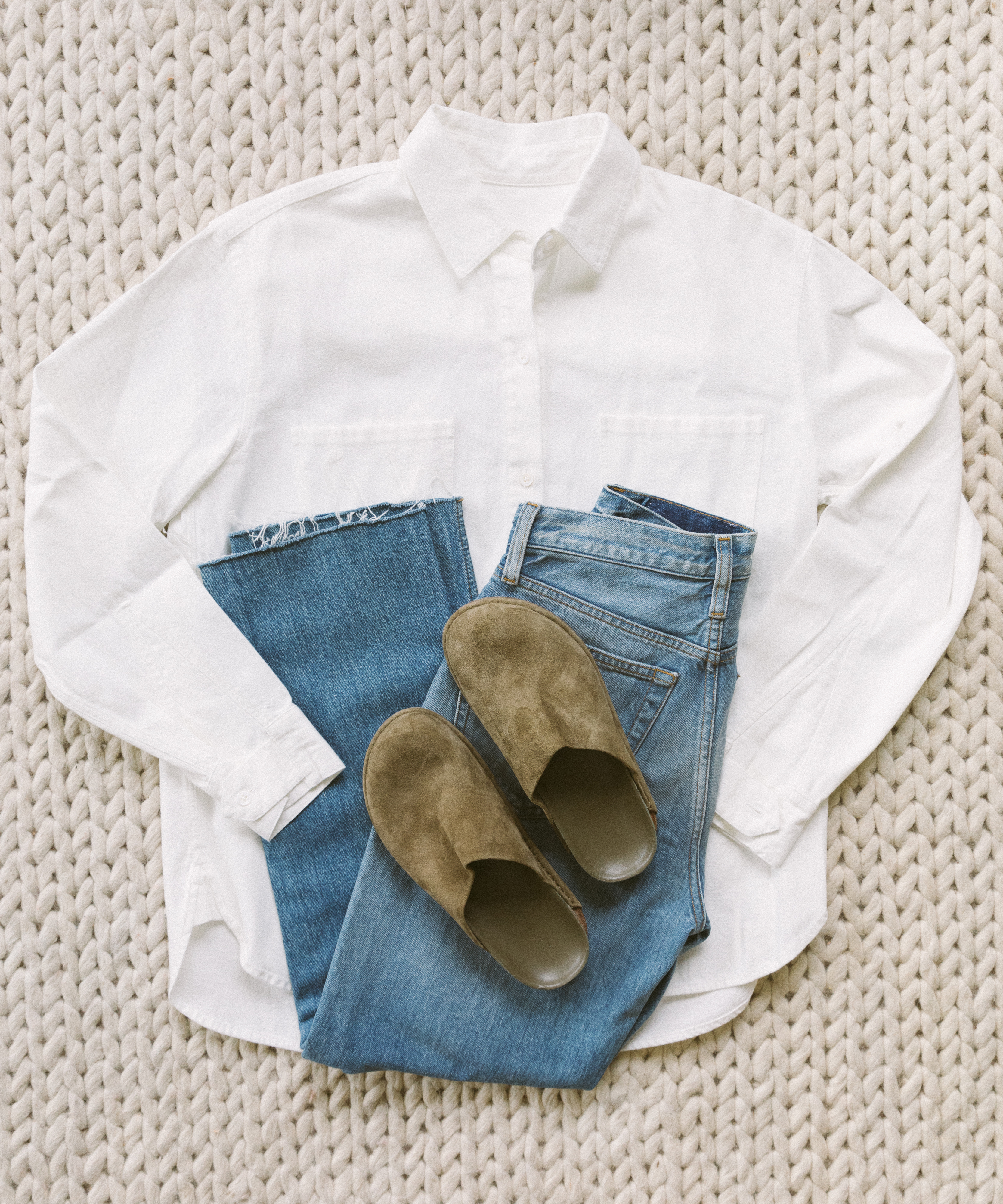 A white button-up shirt, blue jeans, and tan slip-on shoes—essentials for any mountain packing list—are neatly arranged on a textured, cream-colored knit surface.