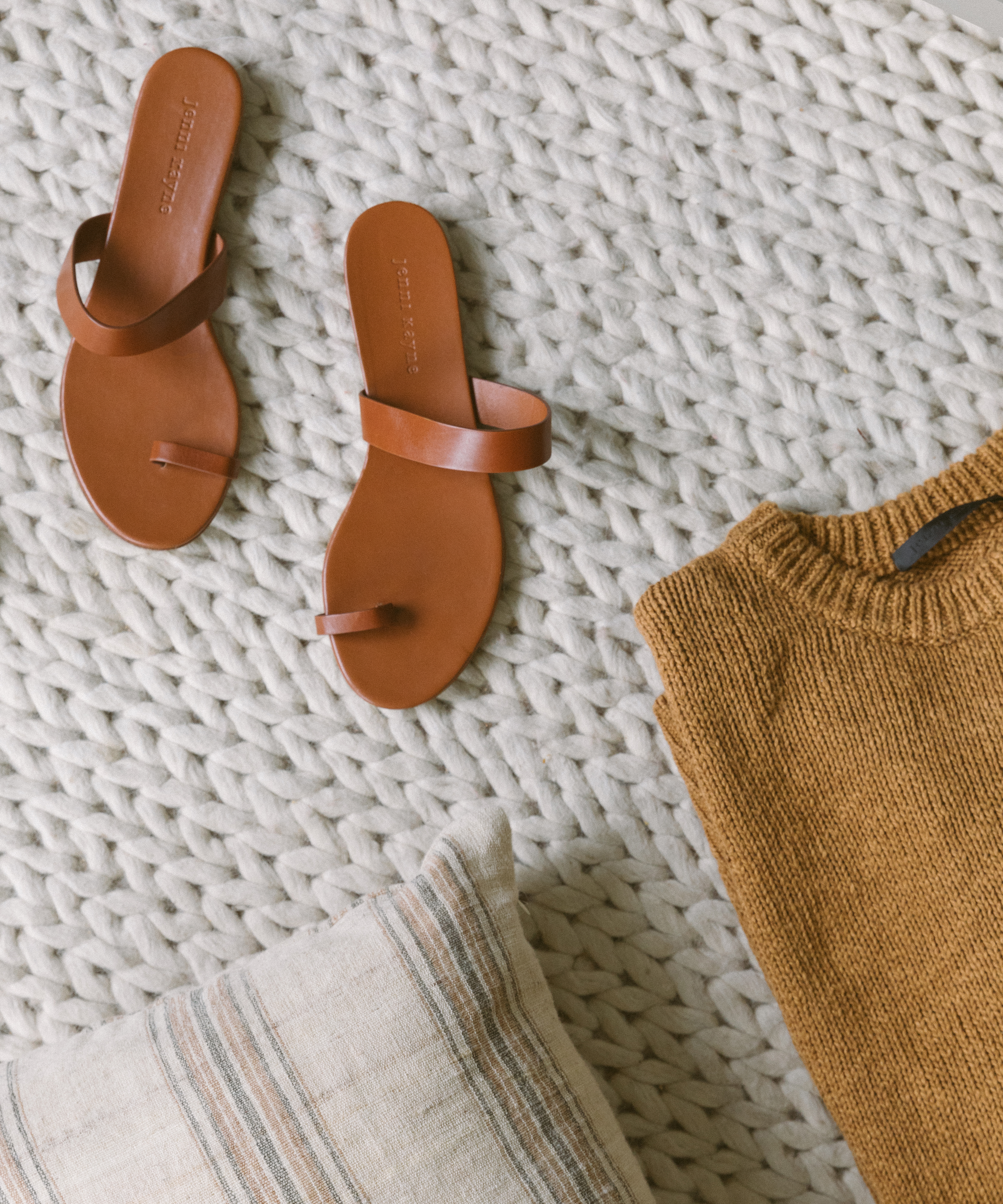 Brown leather sandals, a mustard knit sweater, and a beige-striped pillow arranged on a chunky knit cream blanket—perfect essentials to add to your cozy mountain packing list.