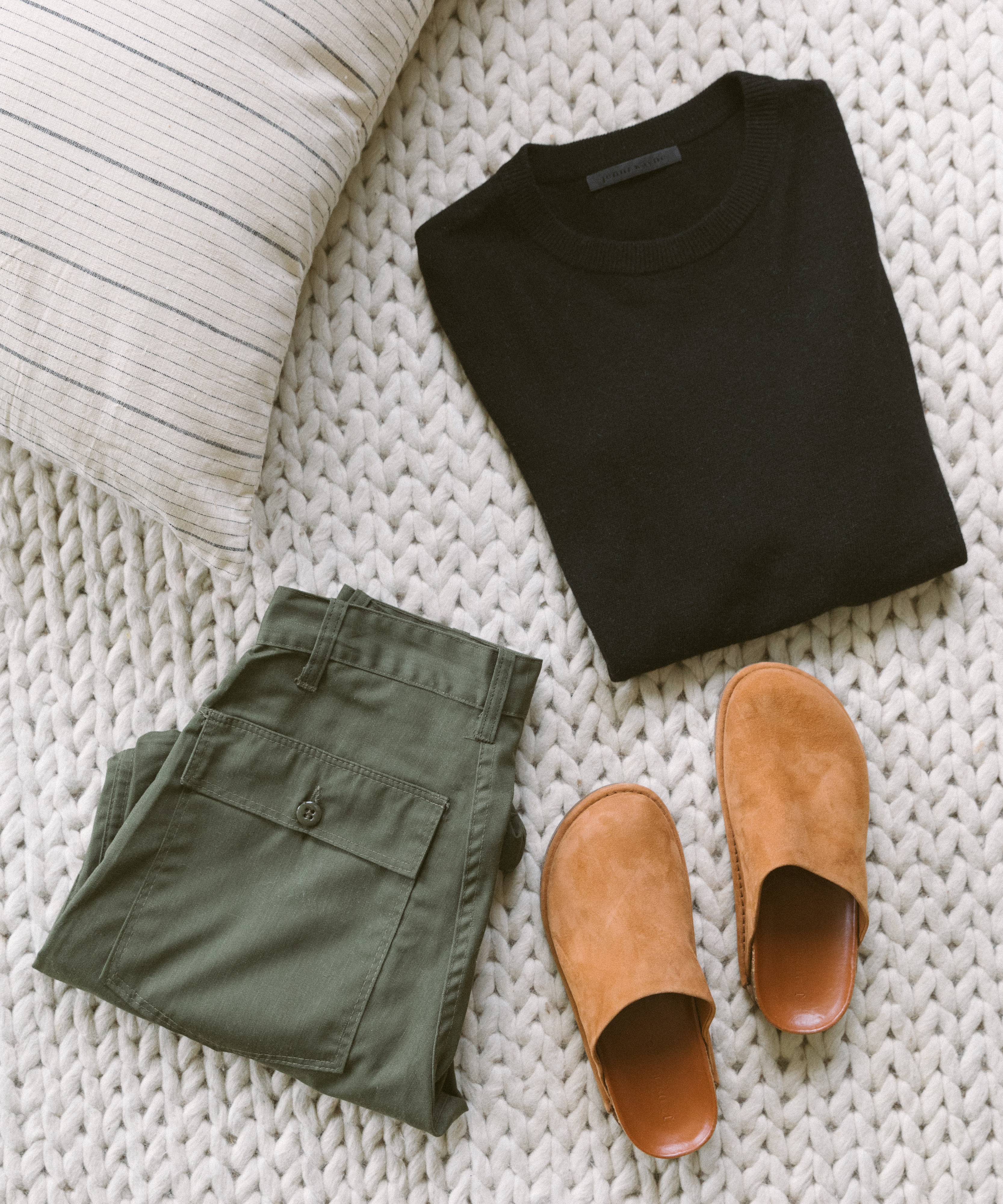 A flat lay of a casual outfit, perfect for your mountain packing list: olive green pants, a folded black sweater, brown slip-on shoes, and a striped pillow on a chunky knit cream blanket.