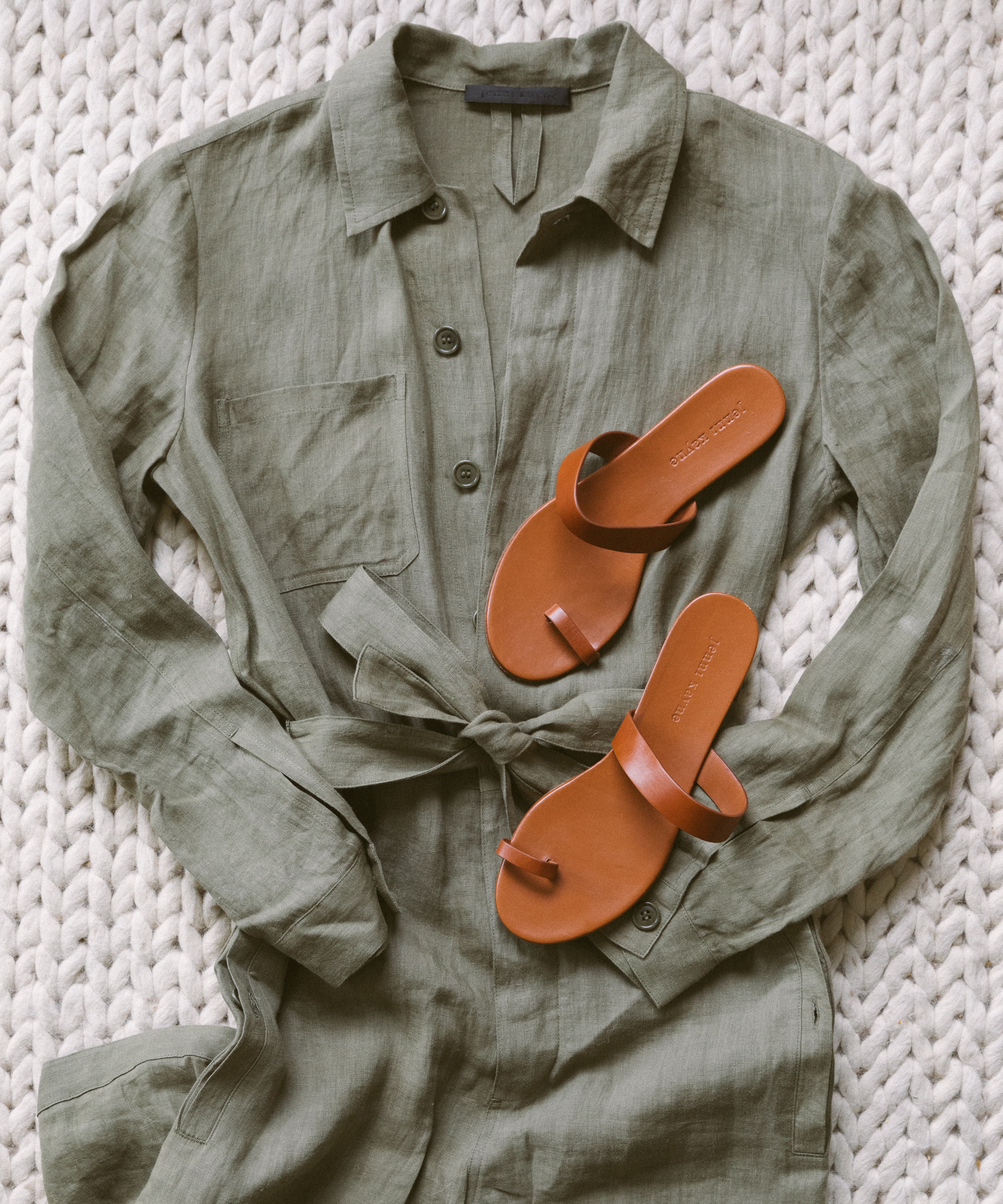 An olive green button-up jumpsuit with a waist tie is laid flat on a chunky knit surface, with a pair of tan leather sandals on top—essentials that earn their spot on any stylish mountain packing list.