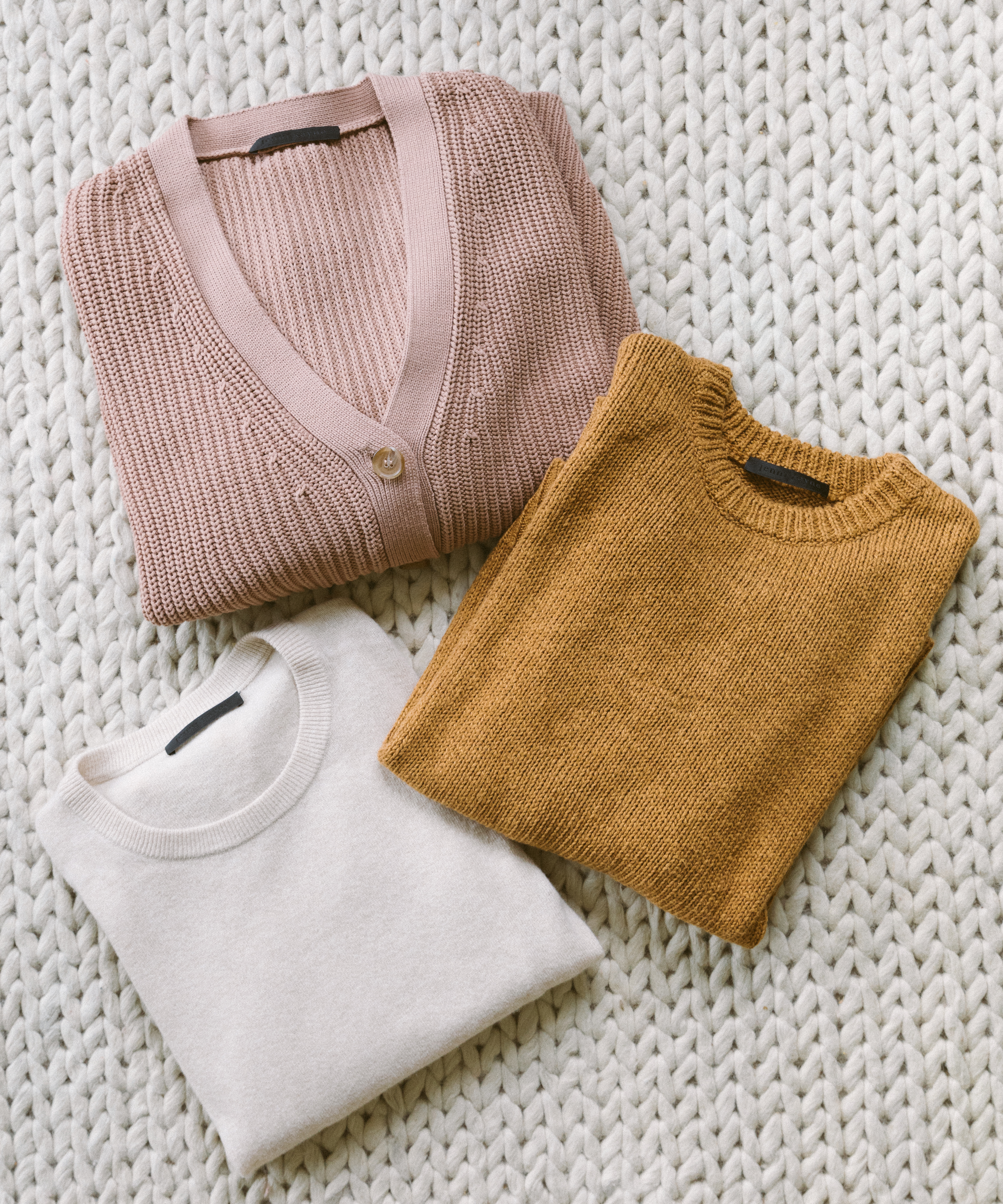 Three neatly folded sweaters—a pink cardigan, a mustard yellow pullover, and an off-white pullover—are arranged on a chunky knit white blanket, perfect additions to your mountain packing list.