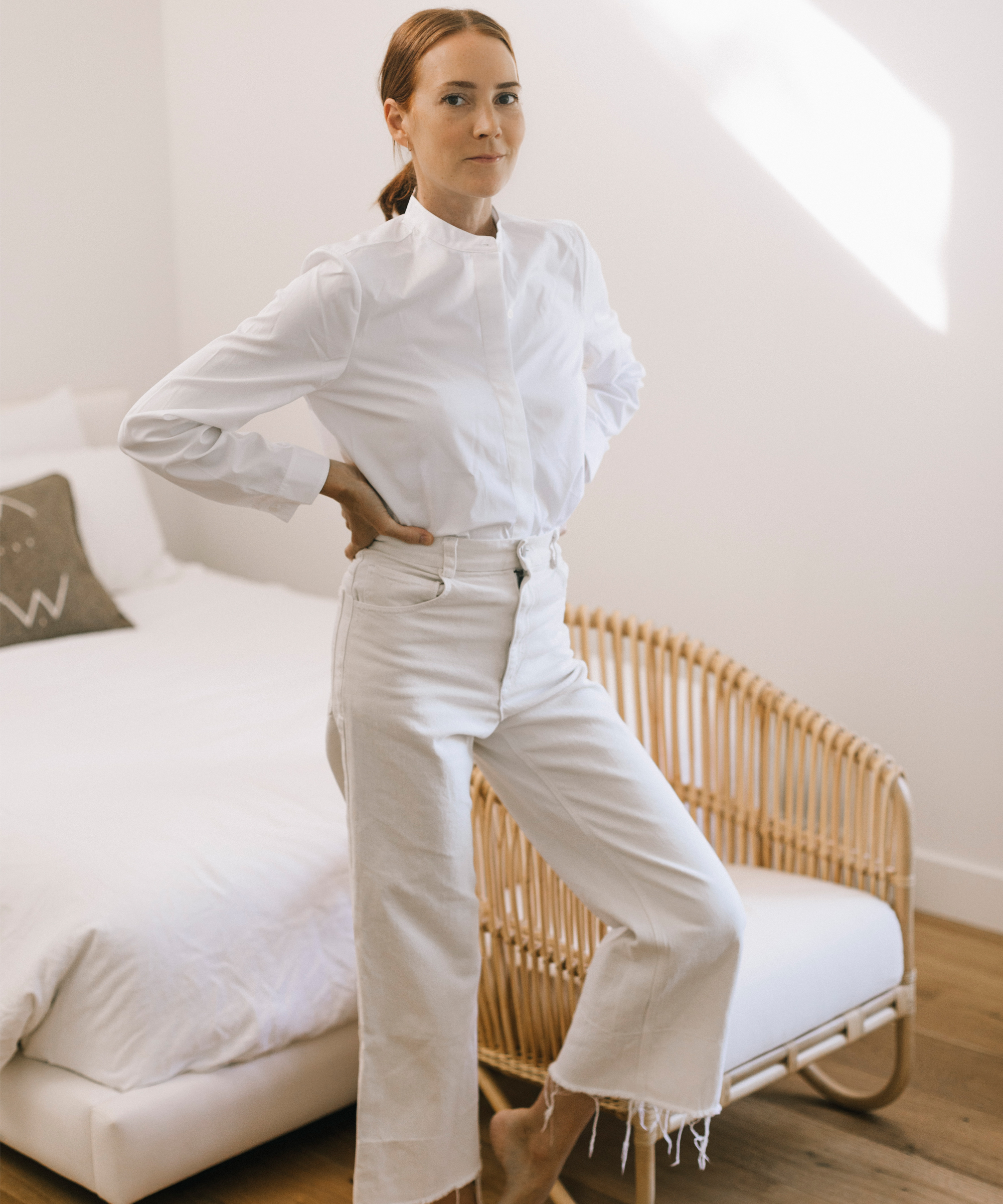 Woman wear button up white shirt and white pants standing in front of a bed.