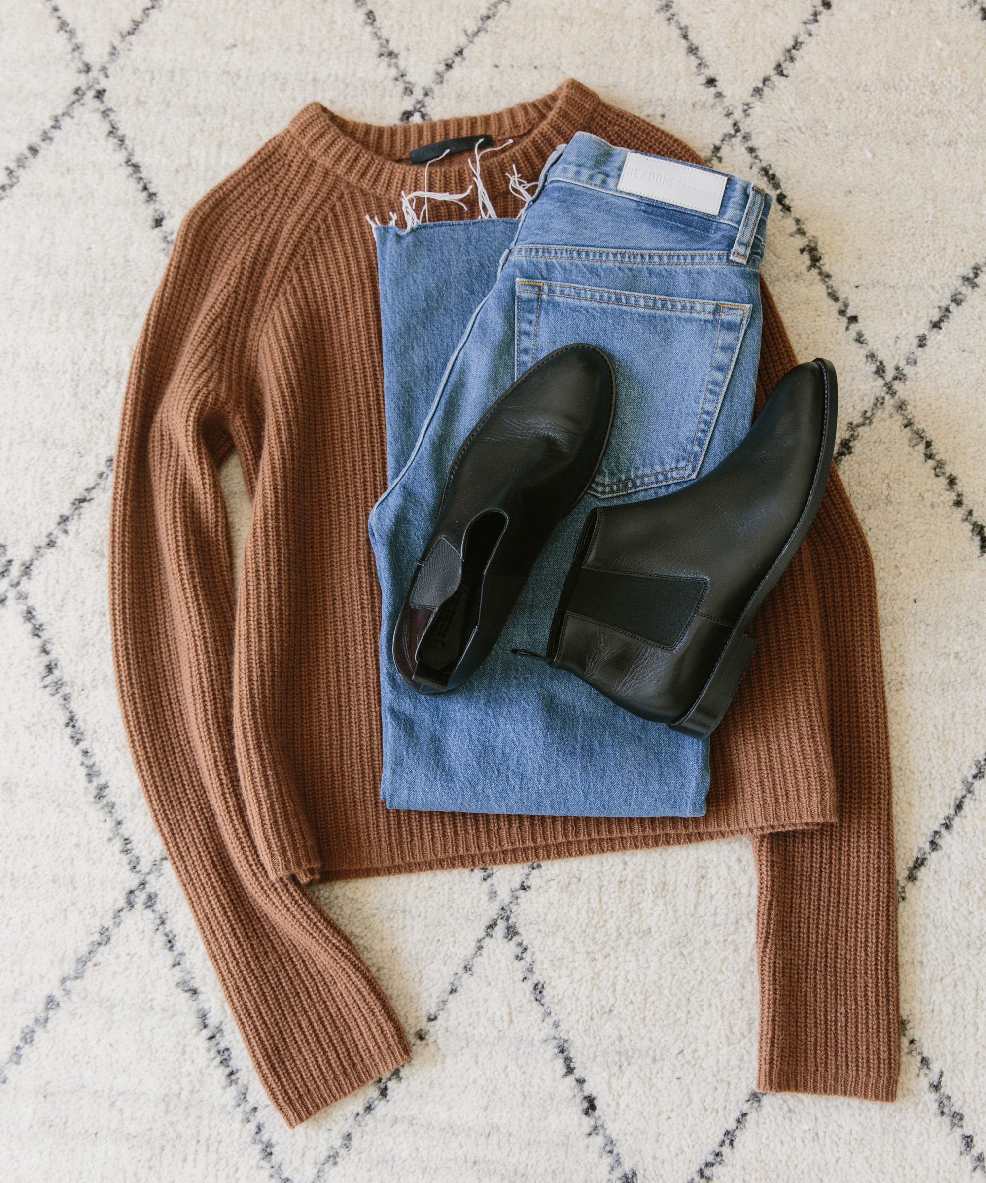 A flat lay of a brown ribbed sweater, blue jeans, and a pair of best chelsea boots arranged on a cream and gray patterned rug.