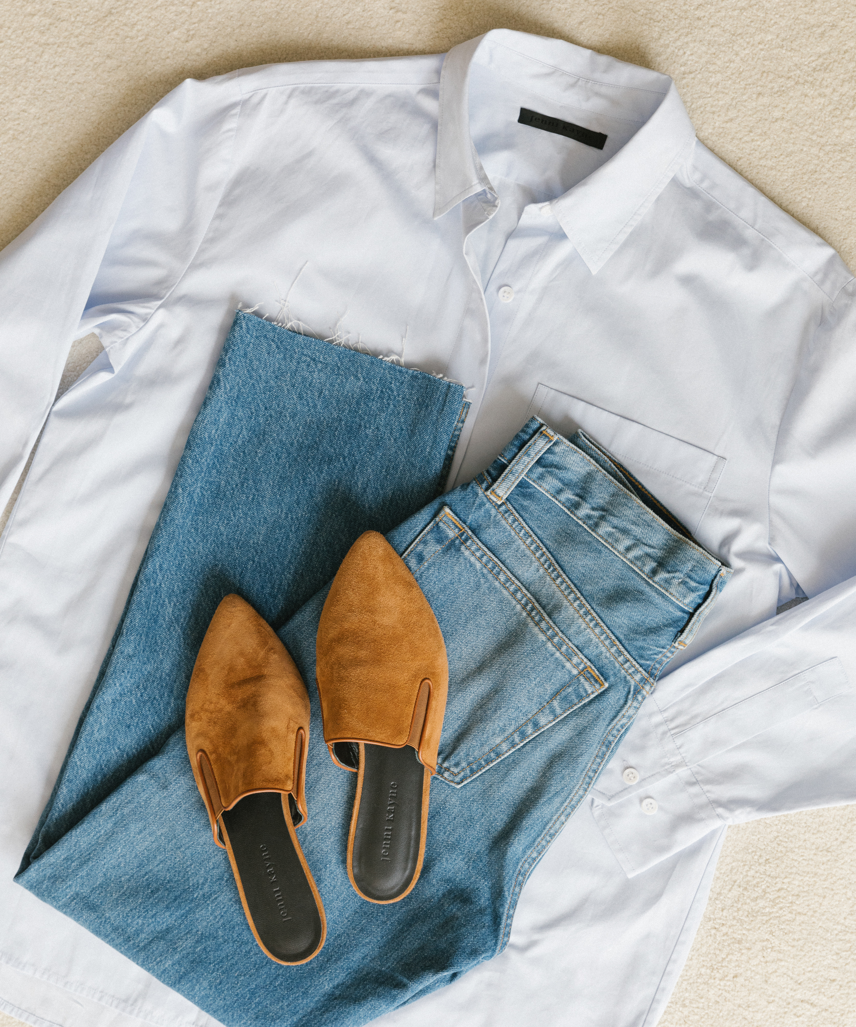 A neatly folded white button-up shirt, blue jeans, and tan suede pointed-toe slip-on shoes are arranged on a light-colored surface—perfect pieces for effortless fall style.