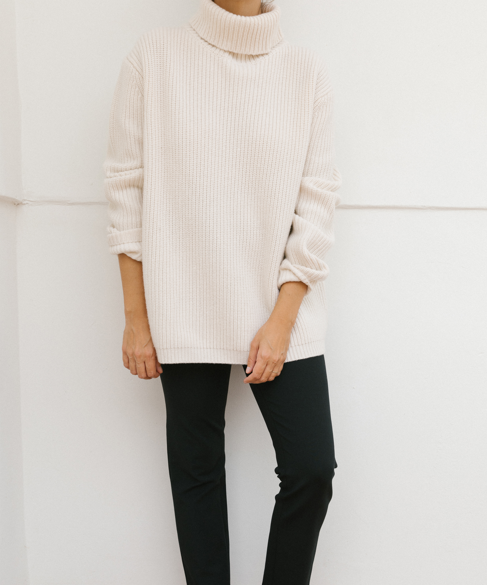 A person stands against a white wall wearing a cream-colored, ribbed turtleneck sweater with the sleeves pushed up and black pants—a timeless look perfect for fall style. Their face is not visible.