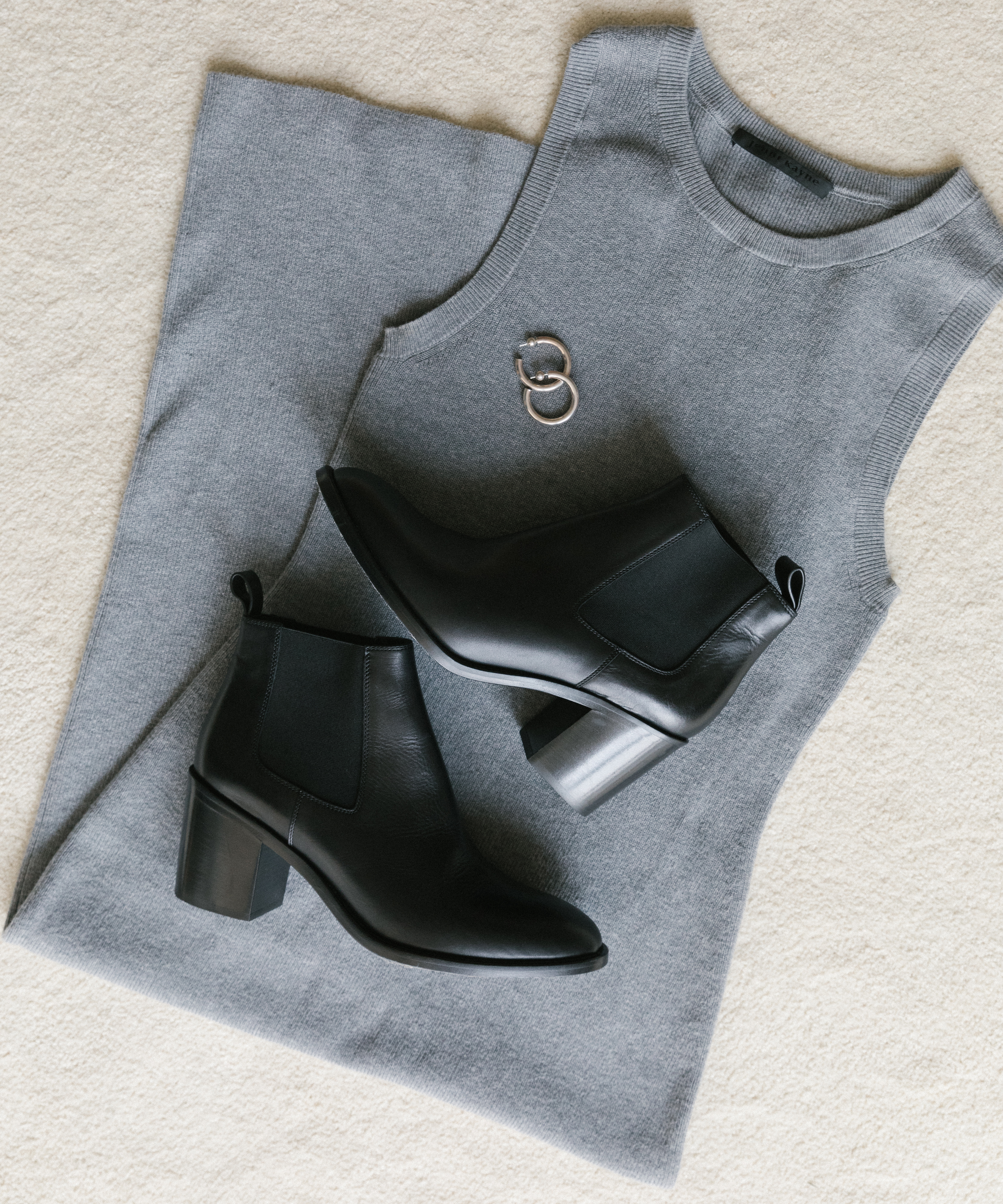 A sleeveless gray knit dress is laid flat on a beige surface, paired with black heeled ankle boots and two silver rings—an effortlessly chic look perfect for fall style.