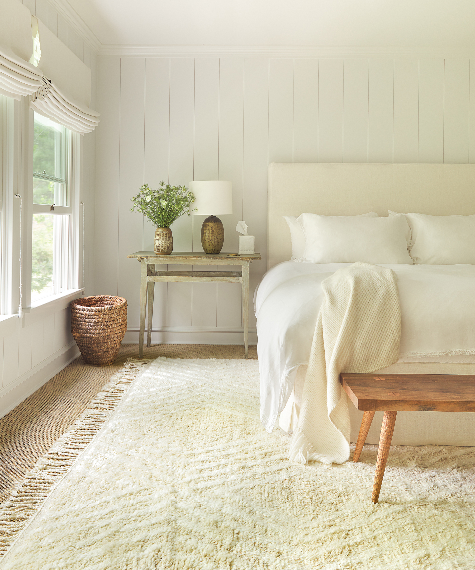 Bright, cozy bedroom with a large bed, white linens, a wooden bench, rustic desk with lamp and vases, woven basket, lulu and georgia rug underfoot, and sunlight streaming through a window with Roman shades.