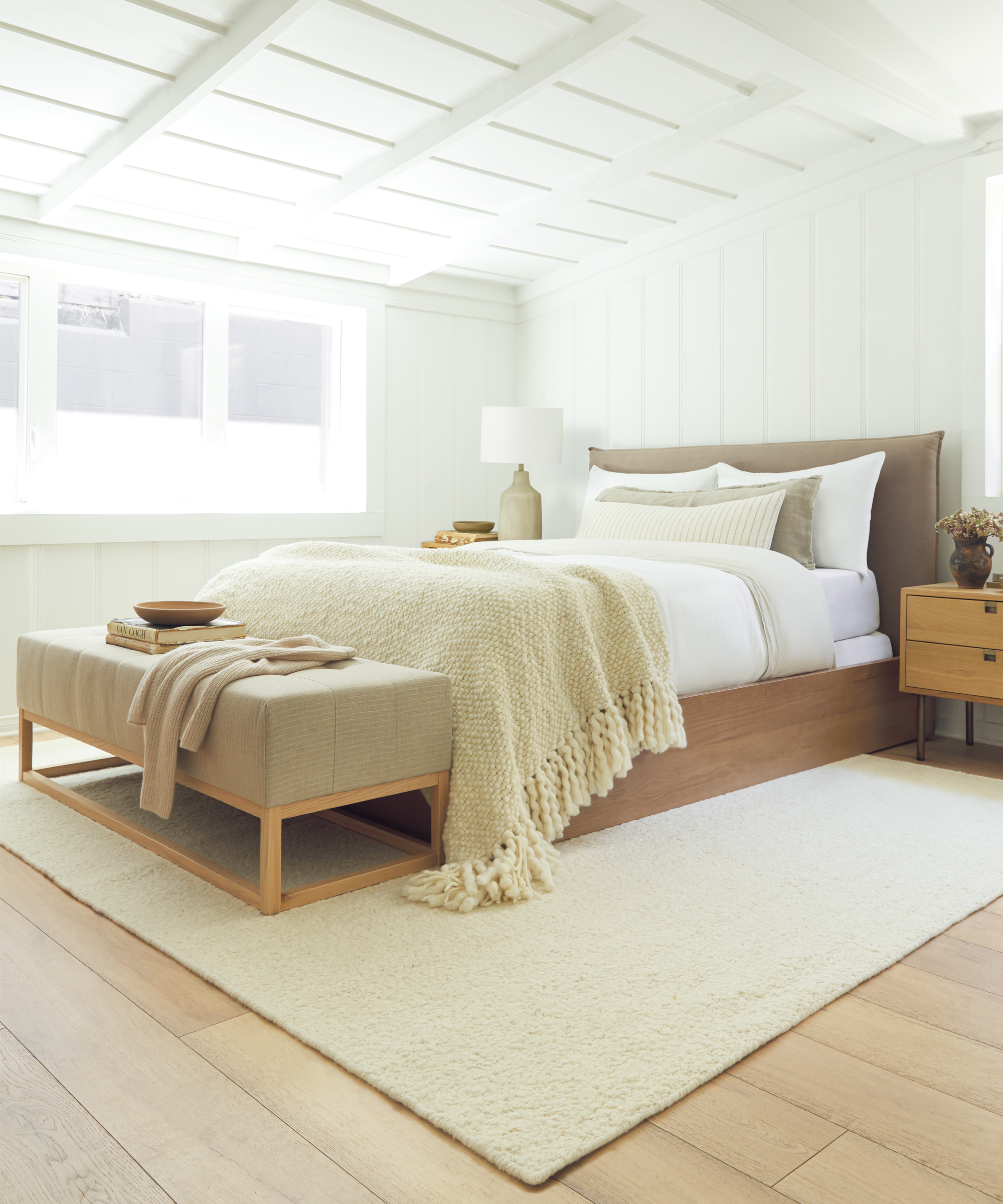 A cozy, modern bedroom with a wooden bed, white bedding, beige throw blanket, cushioned bench, wooden nightstands, a Lulu and Georgia rug, and a large window letting in natural light.