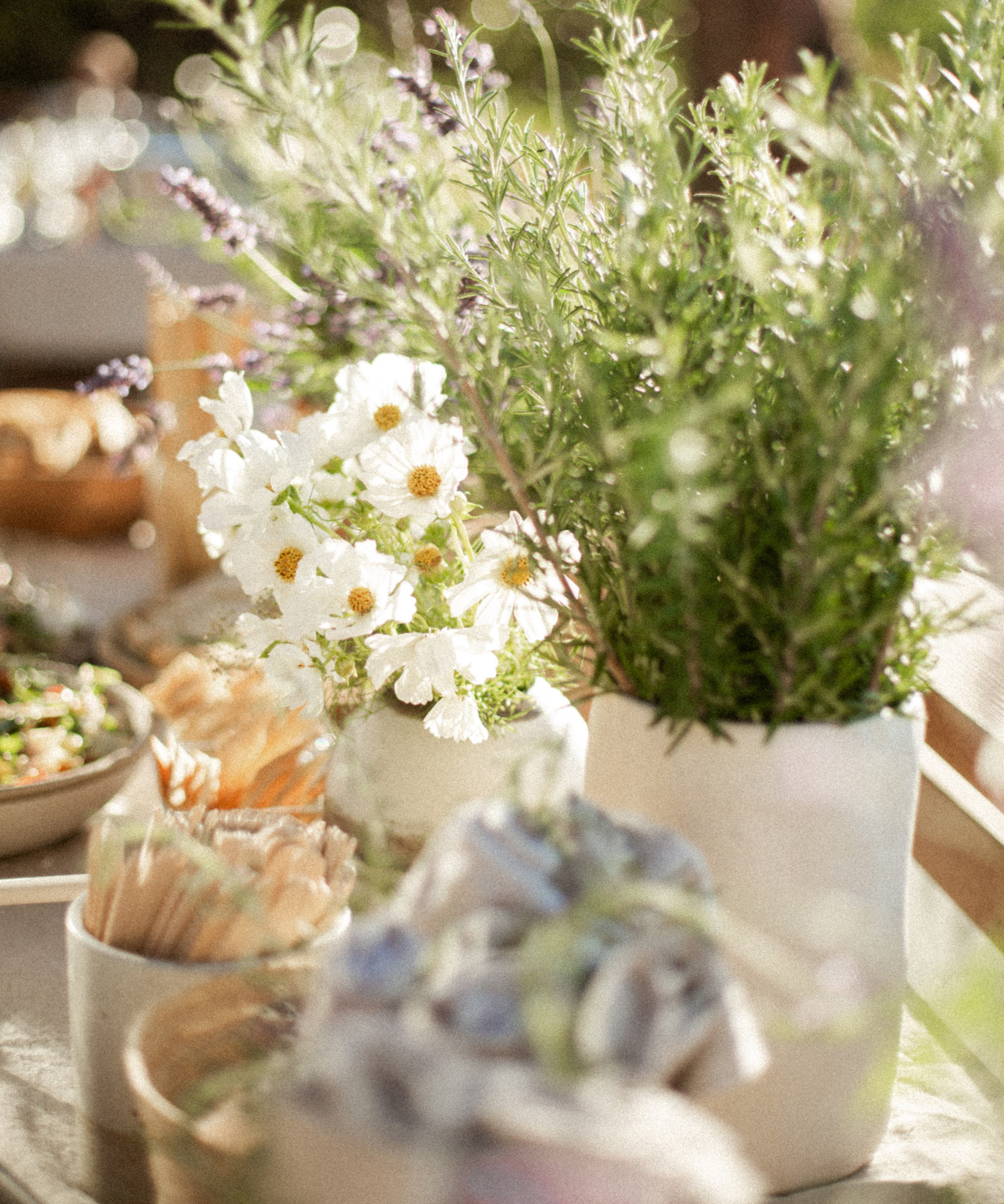 A sunlit outdoor wedding table is decorated with white flowers and green herbs in vases, surrounded by bowls of food and bundles of wooden cutlery, creating a fresh, summery atmosphere.
