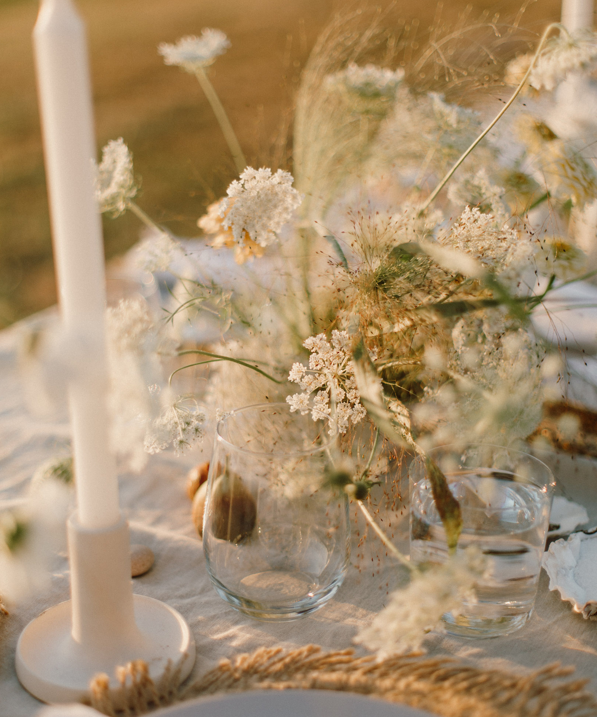 A close-up of a table set for an outdoor wedding with a white tablecloth, white taper candles, clear drinking glasses, and a centerpiece of delicate white wildflowers in soft, warm sunlight.
