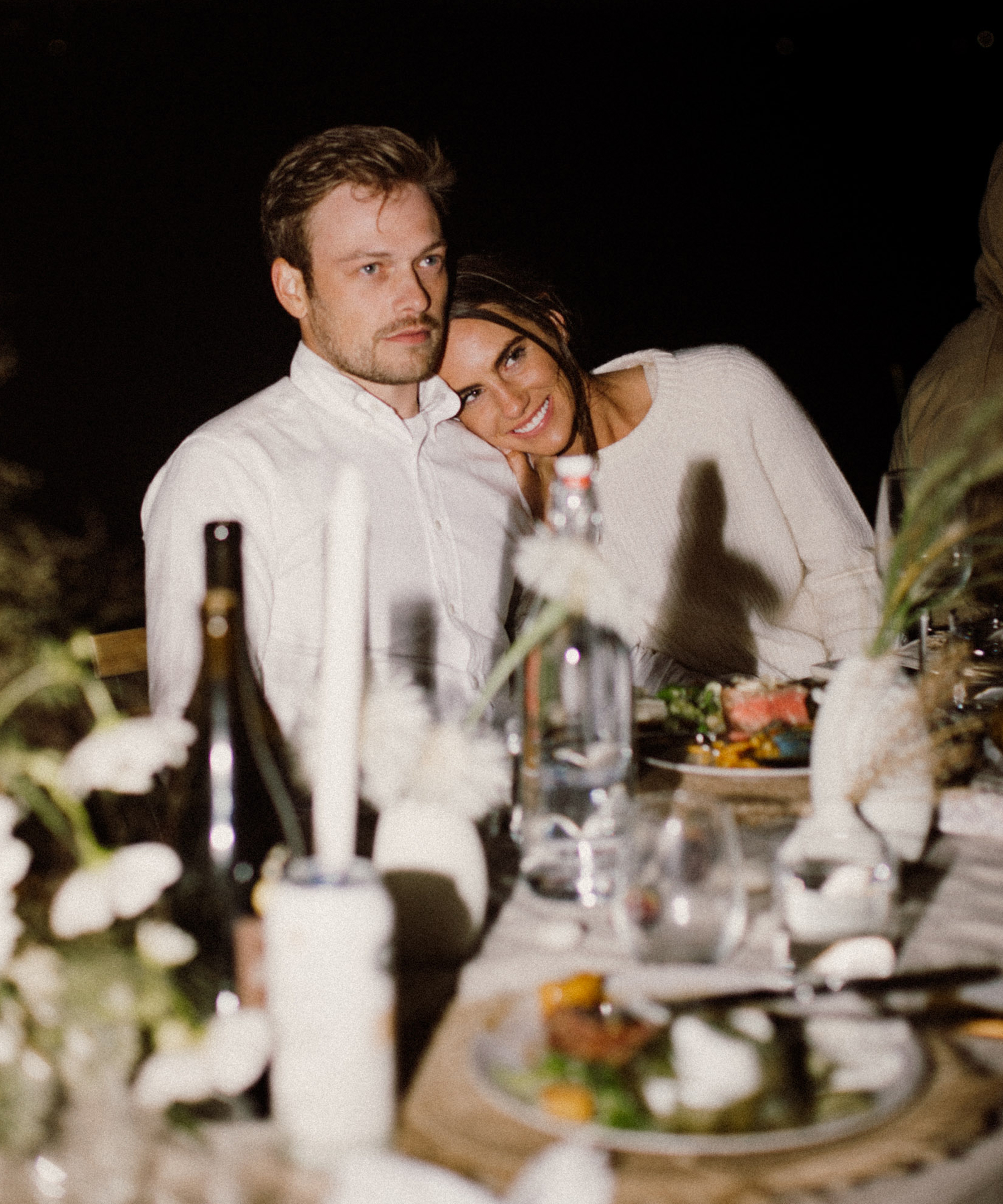 A man and woman sit at a dinner table decorated with flowers and candles during an outdoor wedding. The woman smiles, resting her head on the mans shoulder, while he looks forward with a neutral expression. Plates of food and drinks are on the table.