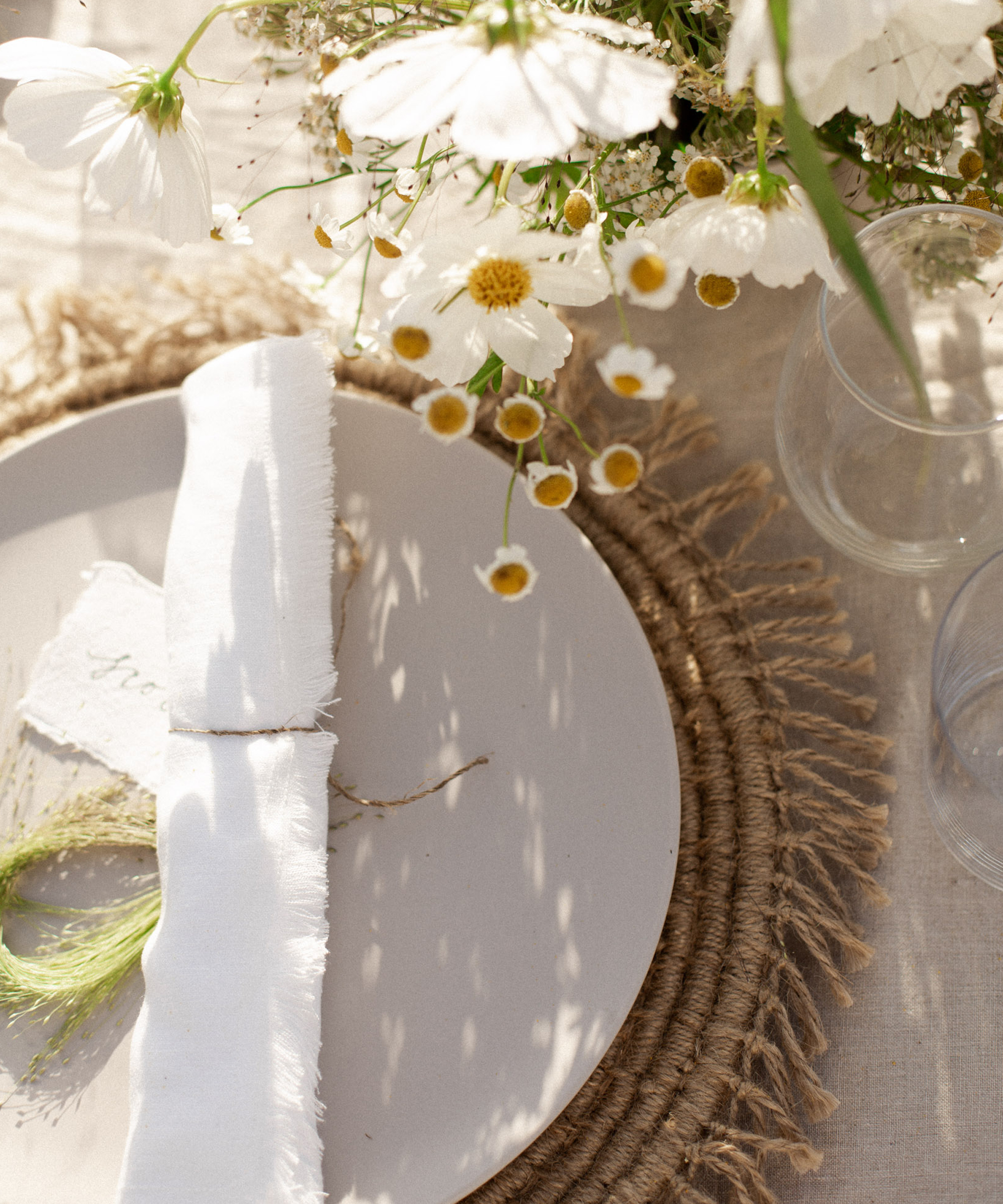 A neutral-toned table setting for an outdoor wedding features a white plate on a woven placemat, a white napkin tied with twine, a sprig of greenery, and wildflowers in a vase. Glassware completes the elegant tablescape.