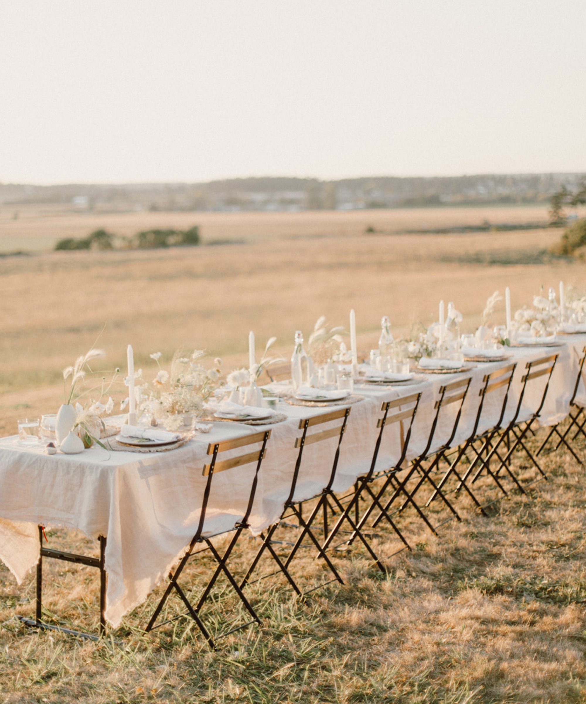 A long outdoor dining table set with white linens, candles, and floral arrangements stands on grass, surrounded by folding chairs—perfect for an outdoor wedding overlooking a sunlit open field and distant horizon.