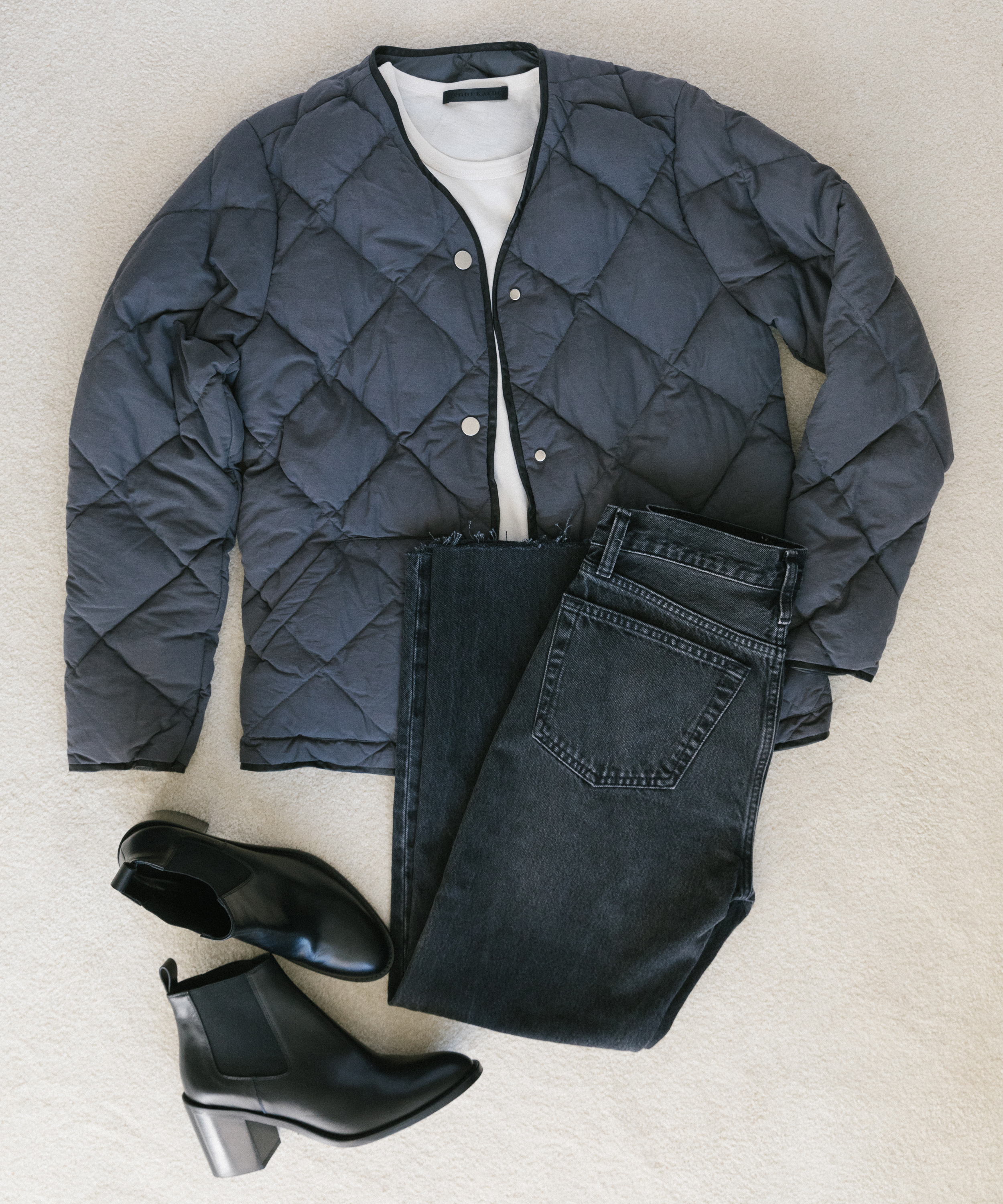 A flat lay of a quilted black jacket, folded white shirt, dark jeans, and the best fall boots—black heeled ankle boots—arranged on a light-colored surface.