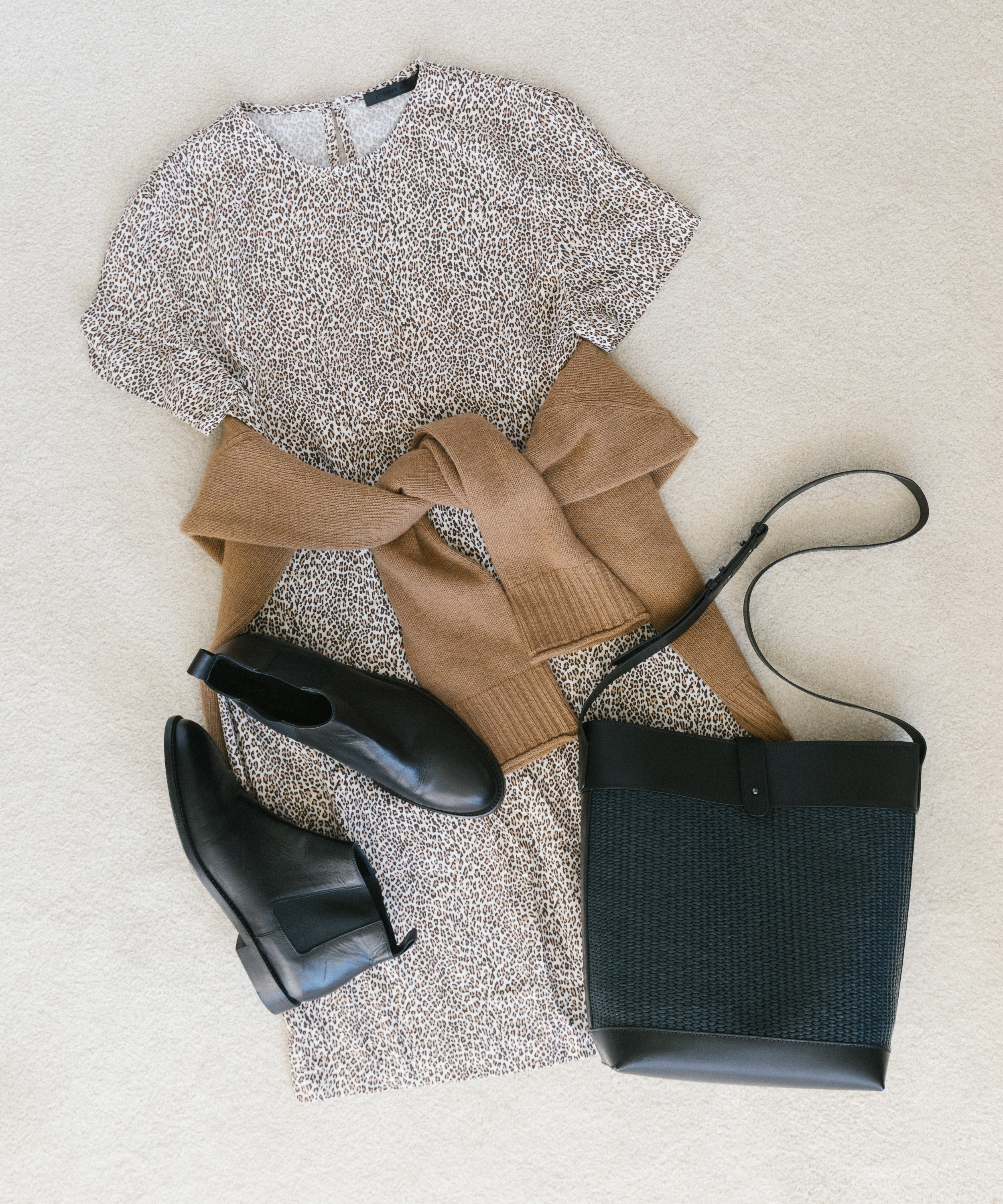A flat lay of a leopard print dress, a tan sweater tied around the waist, black ankle boots—one of the best fall boots—and a black crossbody bag arranged on a light-colored surface.