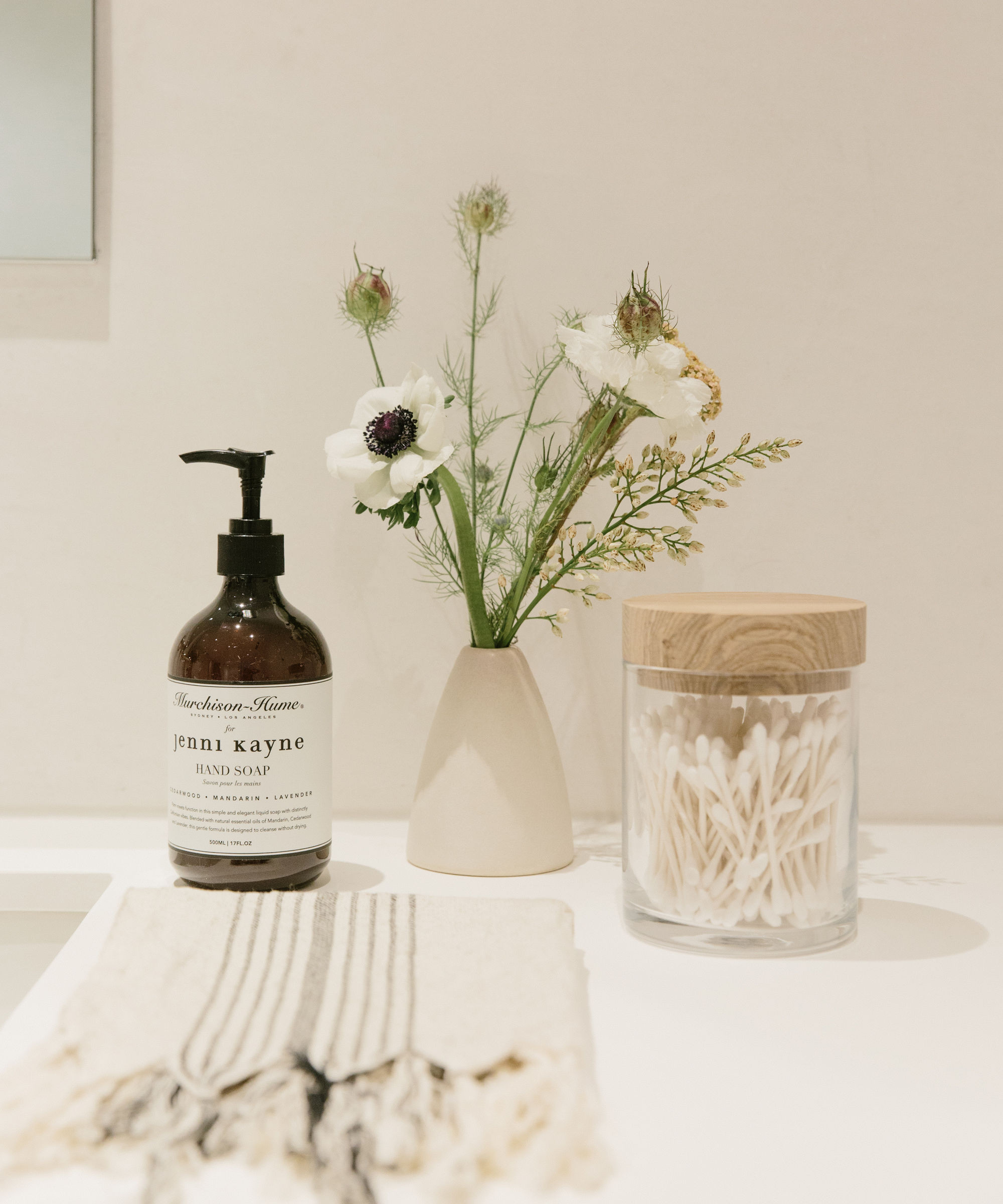 A bathroom counter with a striped hand towel, a bottle of hand soap, a small vase of white flowers, and a glass jar filled with cotton swabs with a wooden lid—perfect for elegant guest room decor in a light, minimal space.
