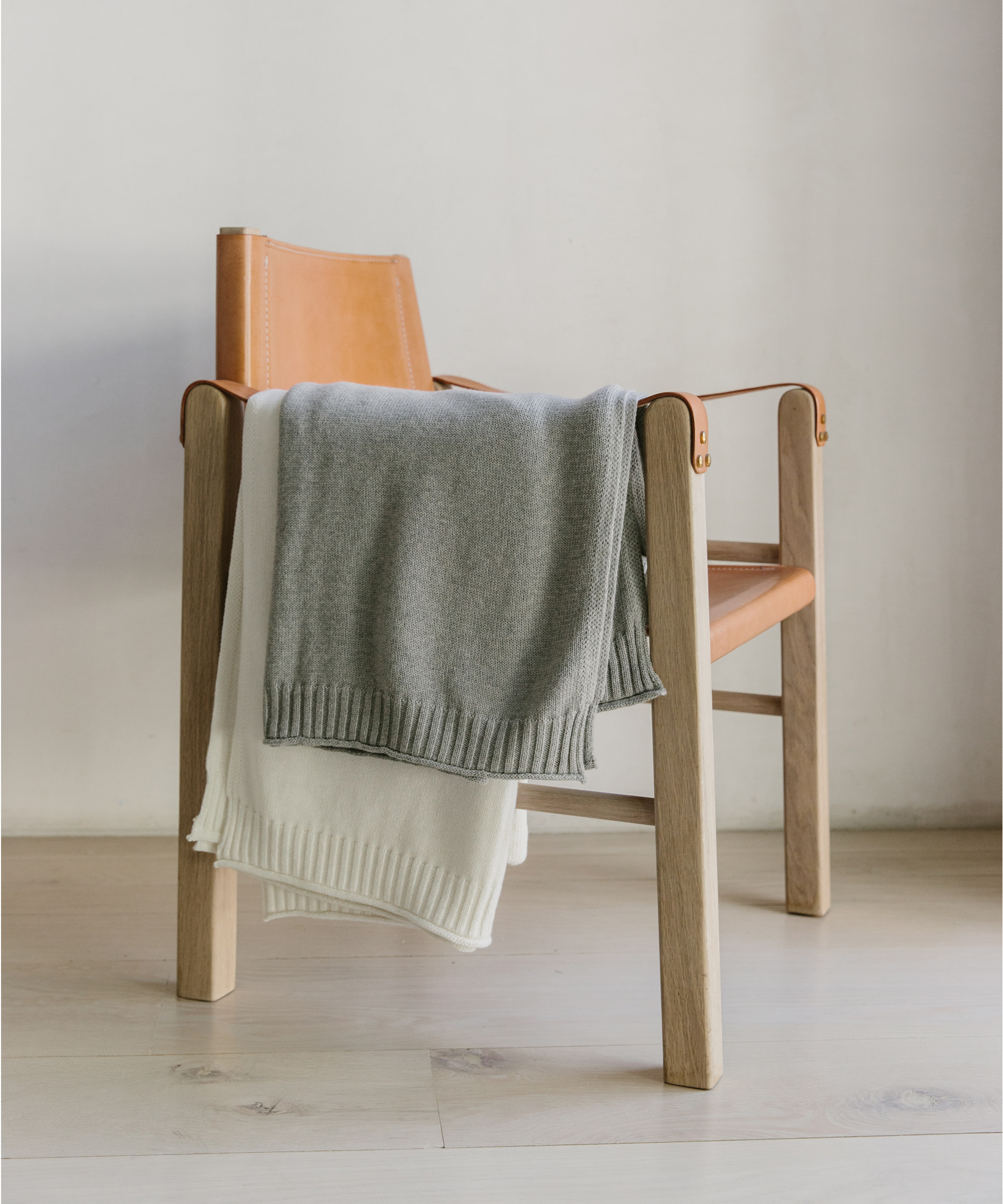 A wooden chair with tan leather seat and backrest, draped with a folded light gray knitted blanket and a folded white knitted blanket—perfectly styled to pair with the best throw pillows—sits on a light wood floor against a plain white wall.
