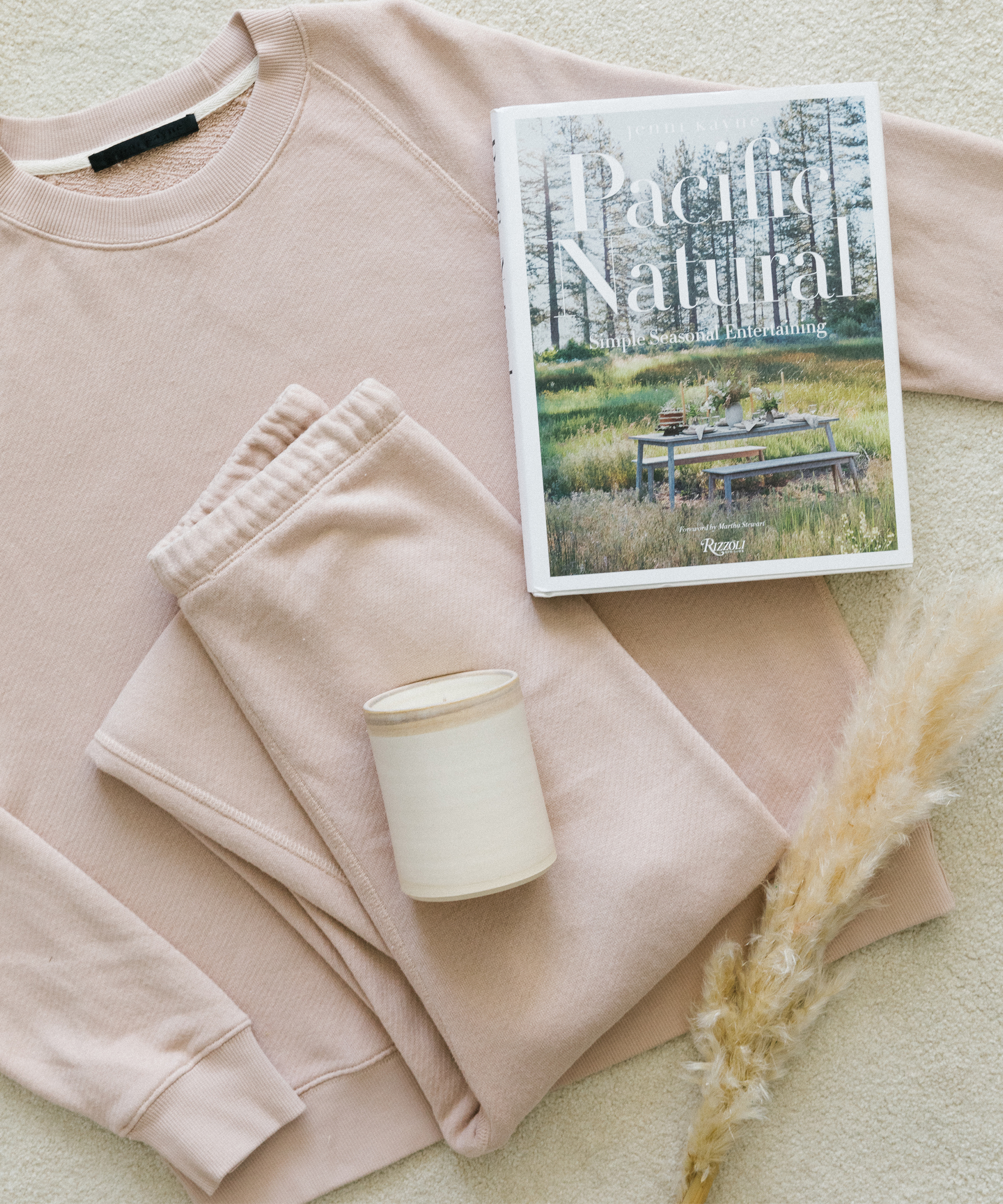 Blush sweatpants and sweatshirt with a ceramic candle, seasonal grasses, and Jenni Kayne's book Pacific Natural sitting on white carpet.
