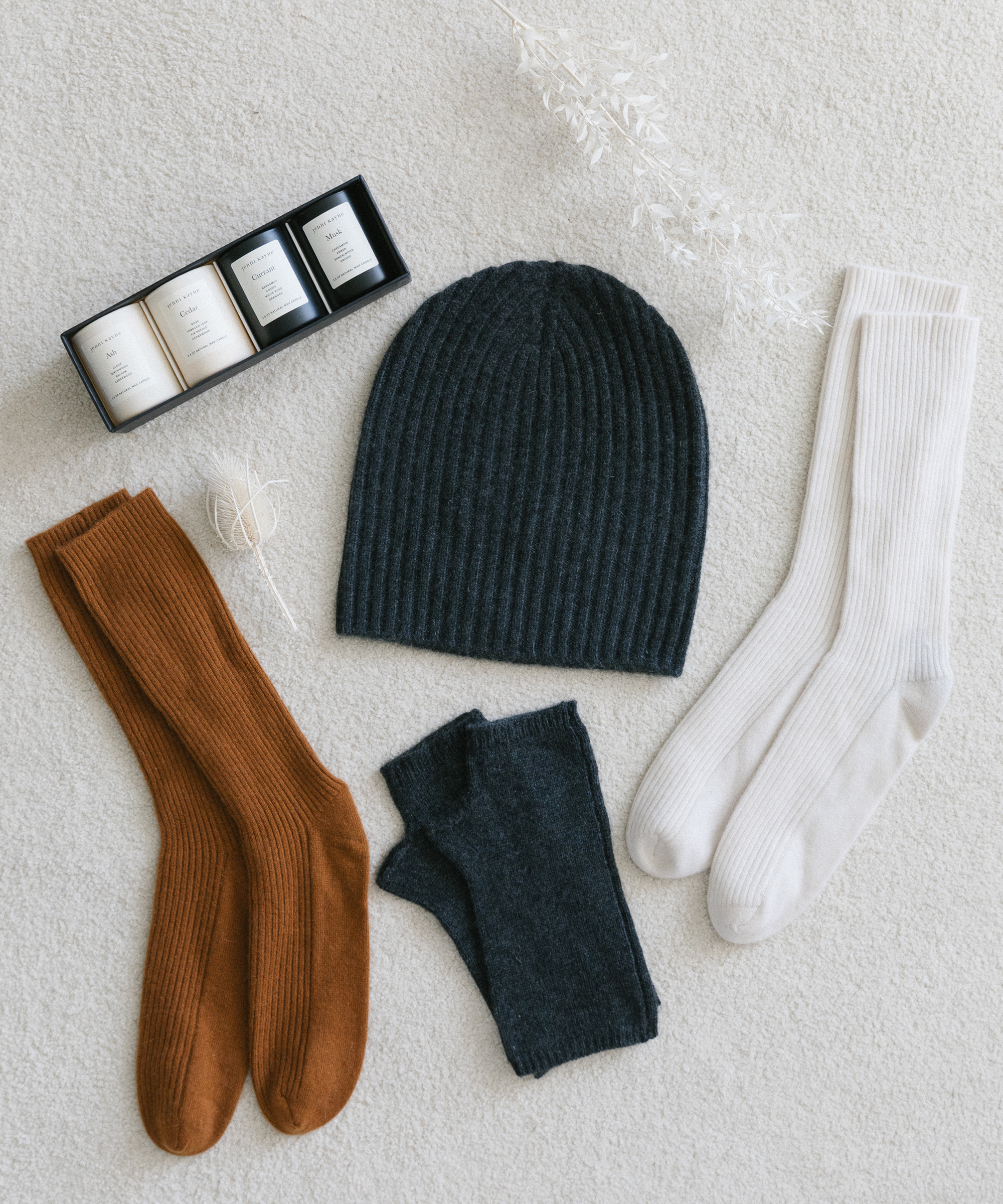 Black cashmere beanie and fingerless gloes, rust socks, and white socks, with votive candle set on top of white carpet.