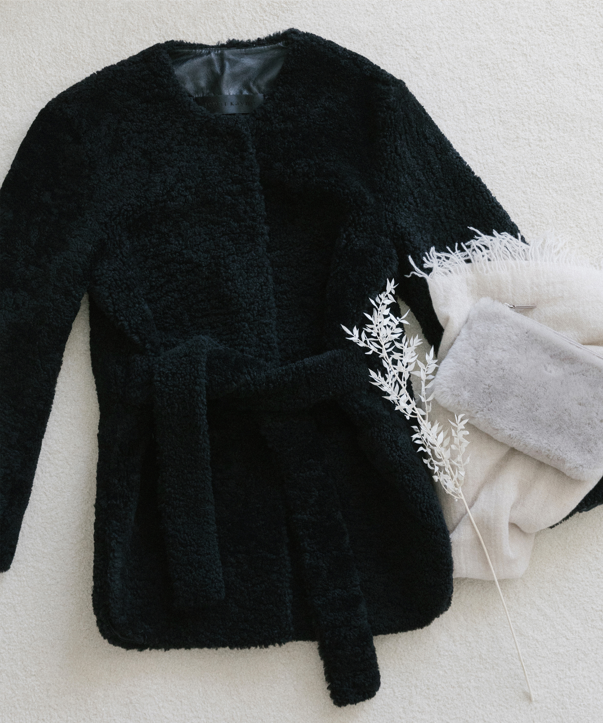 Black shearling coat, grey shearling clutch, and white cashmereshawlon white carpet and seasonal grasses.