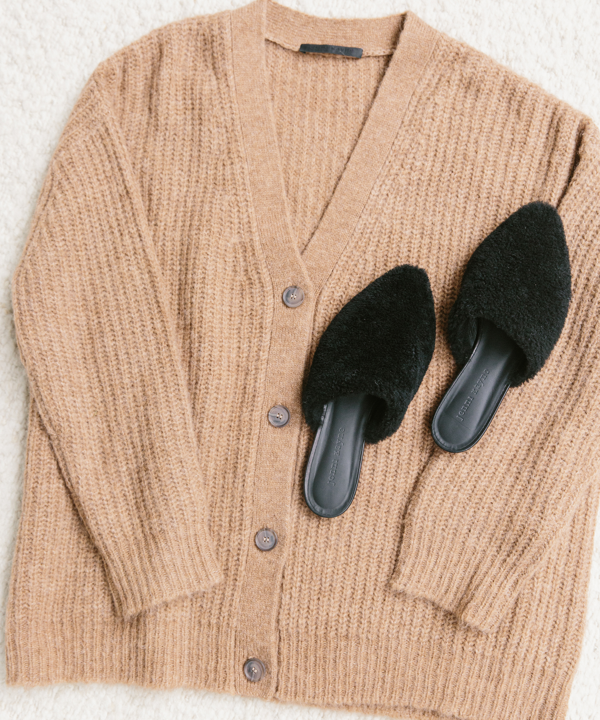 A tan, button-up knit cardigan is laid flat on a light surface, topped with black, fuzzy slip-on slippers—an effortlessly cozy combo perfect for holiday packing.