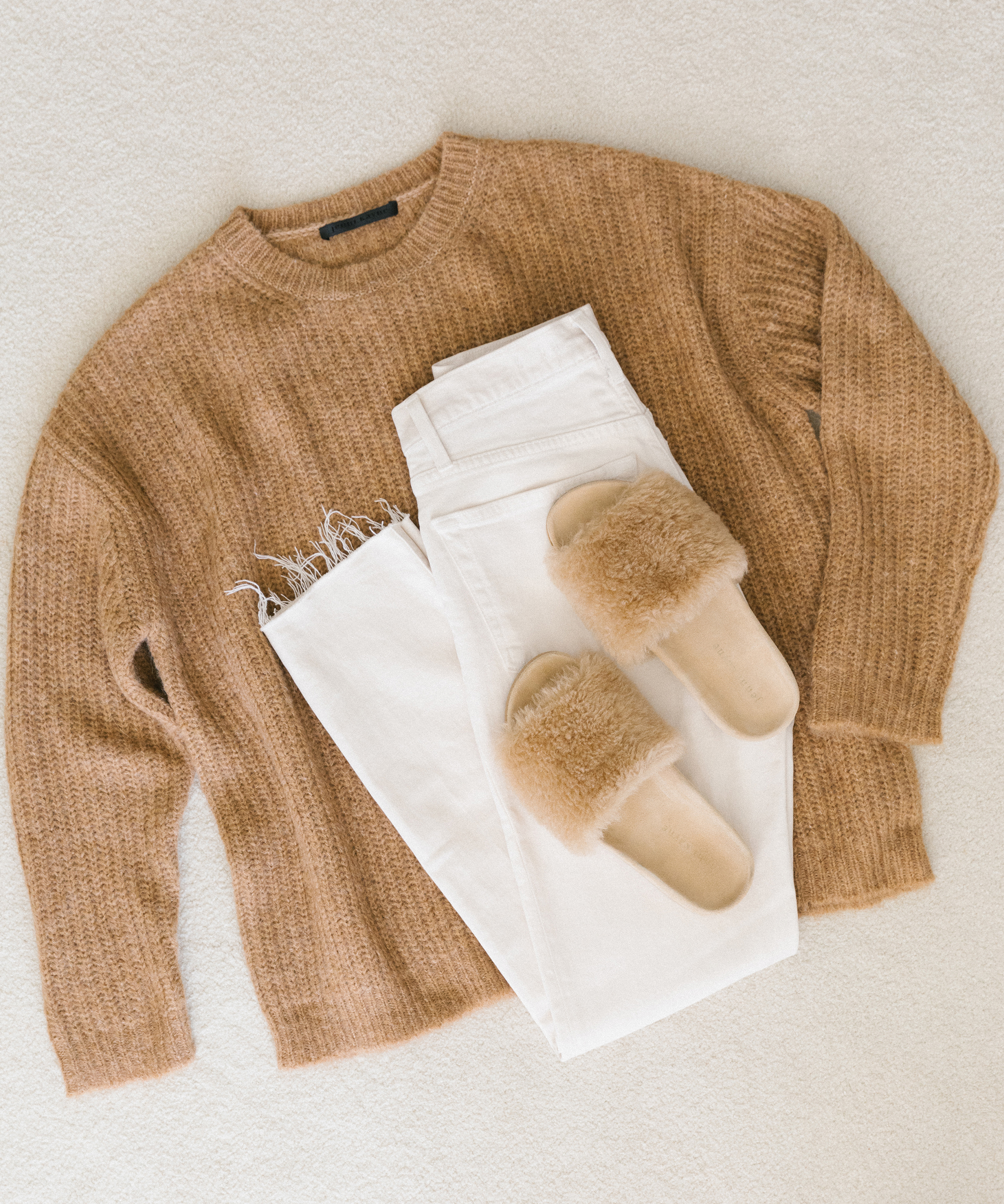 A beige knit sweater, white jeans, and tan fuzzy slide sandals are laid out on a light-colored surface in a flat lay—perfect inspiration for cozy winter outfits.