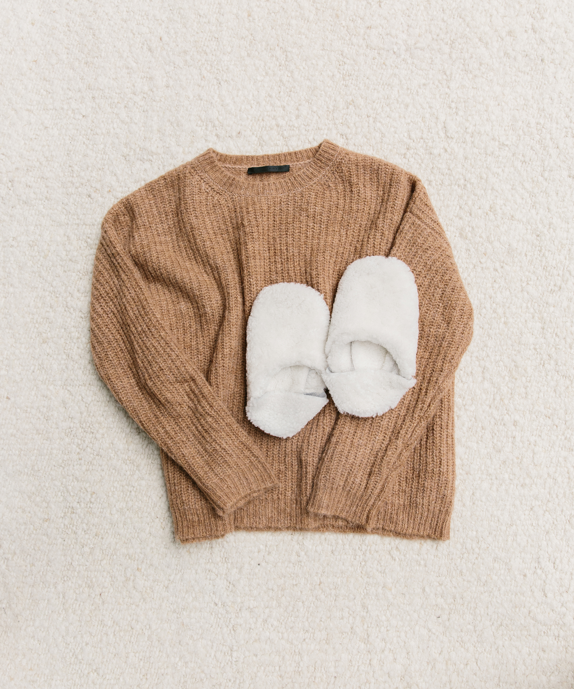 Perfect for holiday packing, this cozy brown knitted sweater with long sleeves lies flat on a soft, cream-colored carpet, with a pair of fuzzy white slippers placed on top.