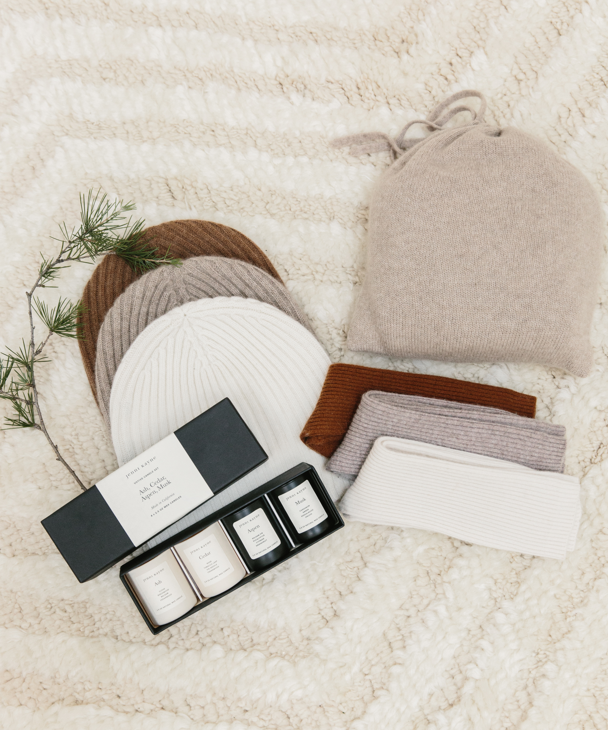 A cozy flat lay featuring ribbed knit hats and headbands in beige, brown, white, and rust, a small pine branch, and a black box of three white scented candles—all perfect as holiday gifts—arranged on a cream textured blanket.