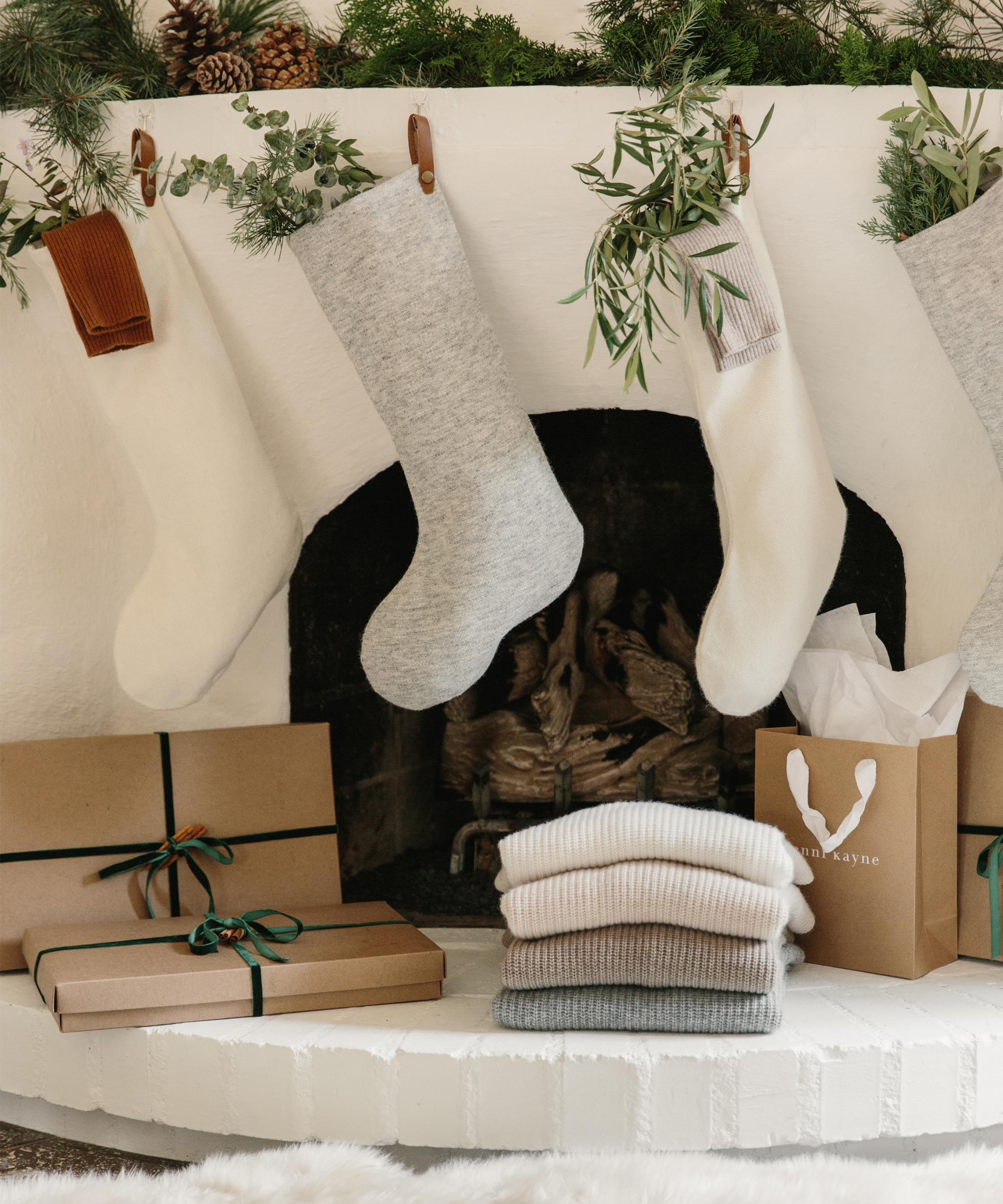 A cozy holiday scene with stockings above a white fireplace, wrapped holiday gifts with green ribbon, a stack of folded sweaters, and a brown gift bag, all decorated with pine branches and greenery.