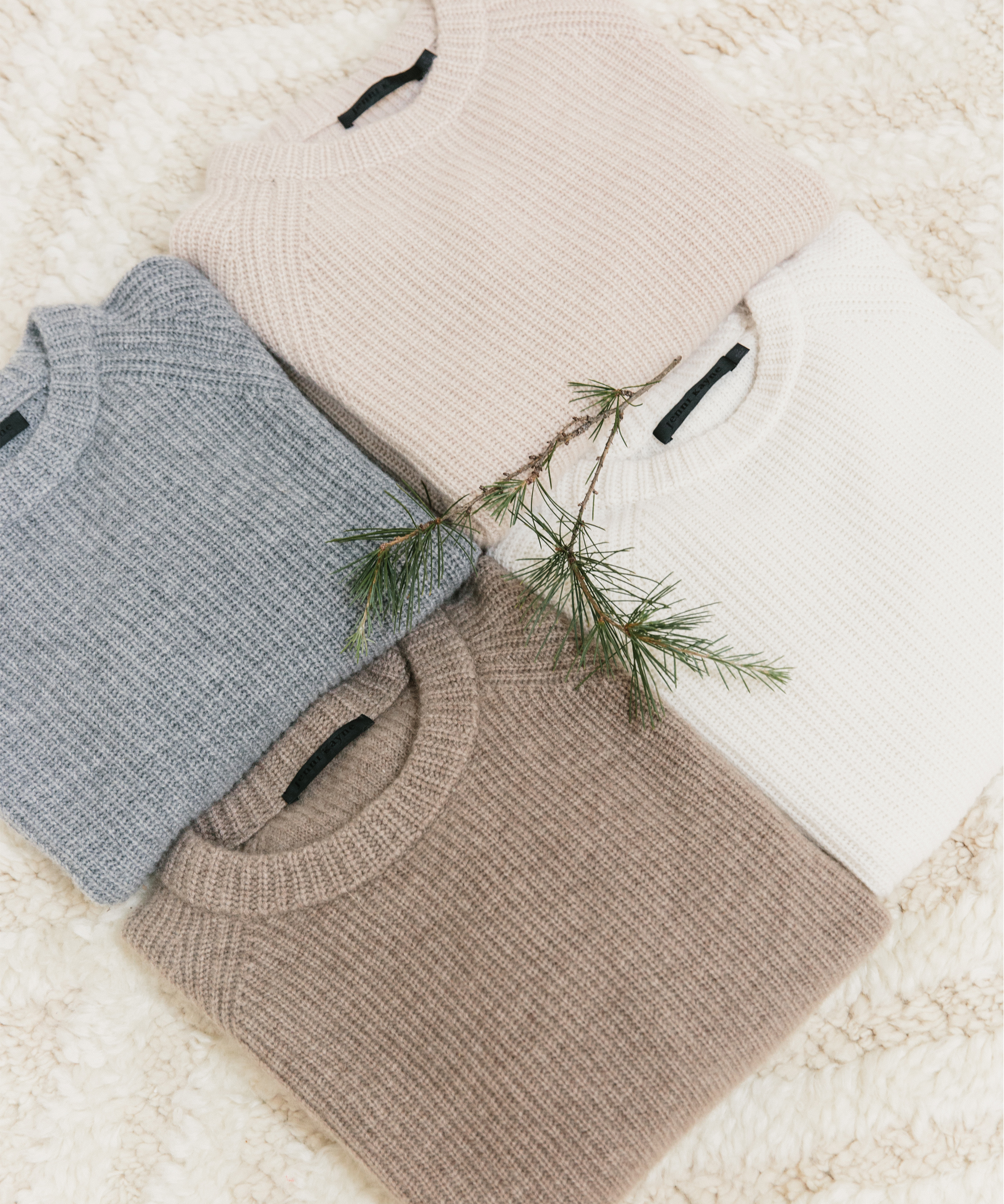 Four folded knit sweaters in soft shades of beige, white, gray, and light taupe are arranged in a square on a textured cream surface. A pine branch rests on the white sweater—perfectly cozy picks for thoughtful holiday gifts.