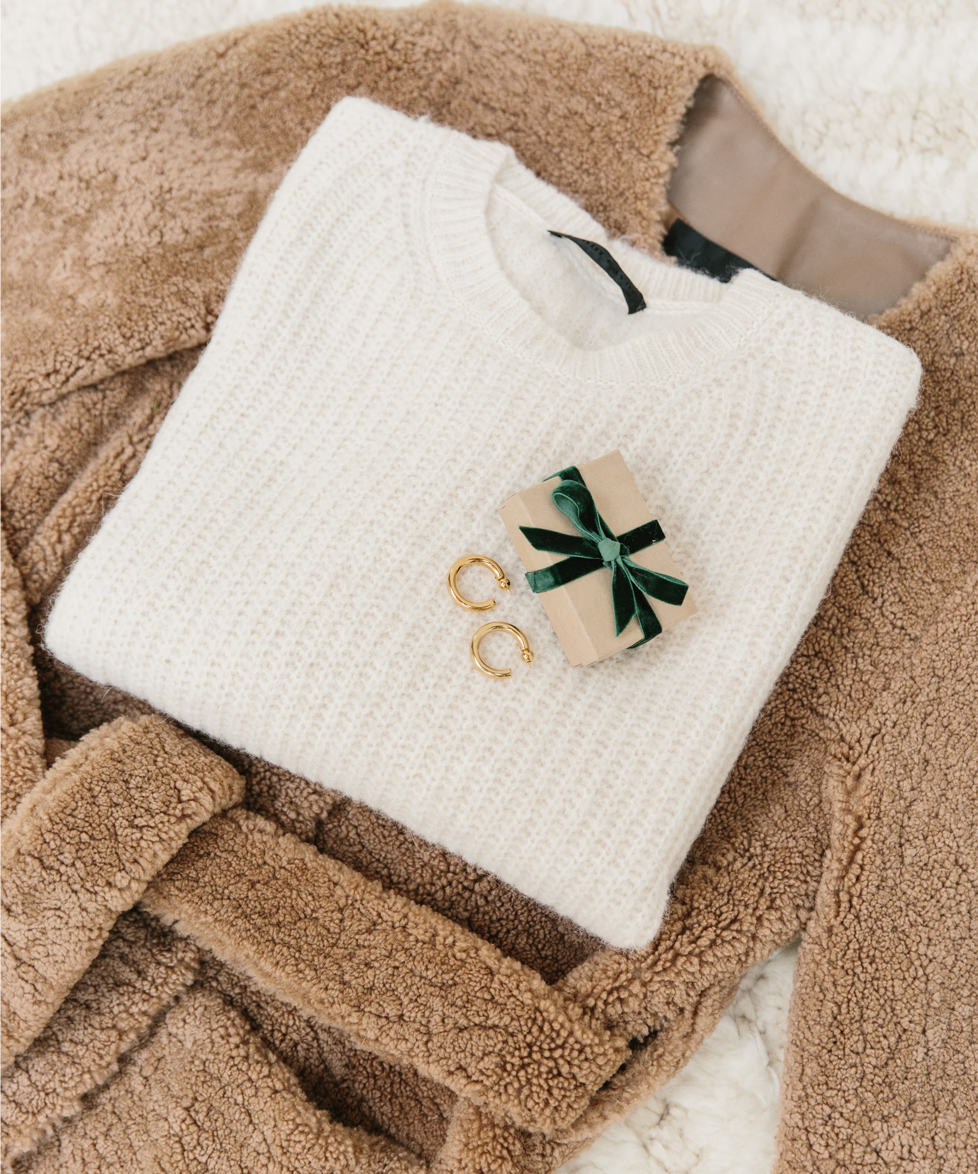 A folded white knit sweater topped with two gold hoop earrings and a small gift box tied with a green ribbon, all arranged on a fluffy brown coat—perfect inspiration for stylish holiday gifts.