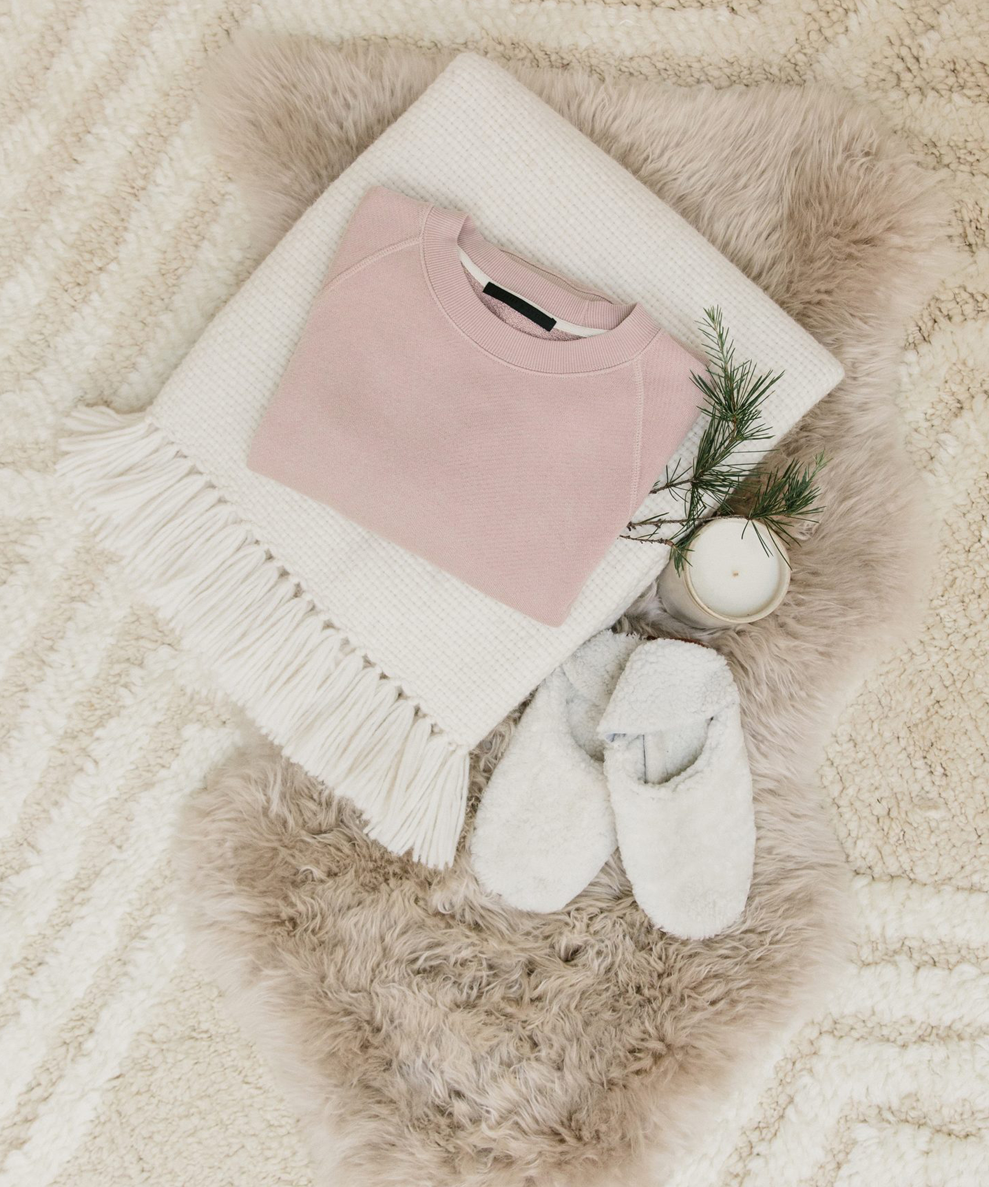 A folded pink sweater, white fringed blanket, gray slippers, a lit candle, and a small pine branch arranged on a beige furry rug over a cream patterned carpet—perfect inspiration for cozy holiday gifts.