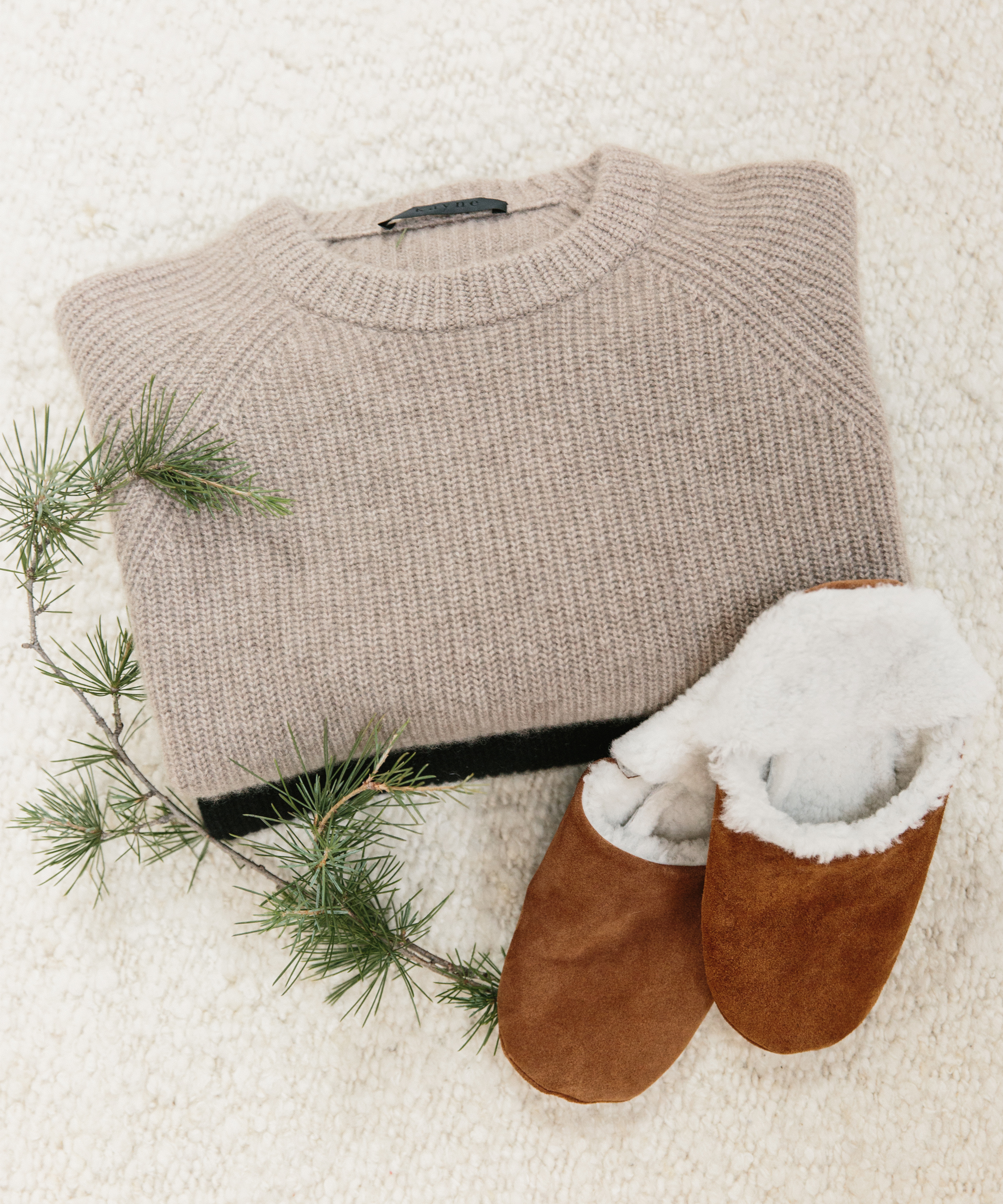 A folded beige knit sweater, a sprig of pine, and a pair of brown suede slippers with white fleece lining rest on a soft, cream-colored surface—perfect inspiration for cozy holiday gifts.