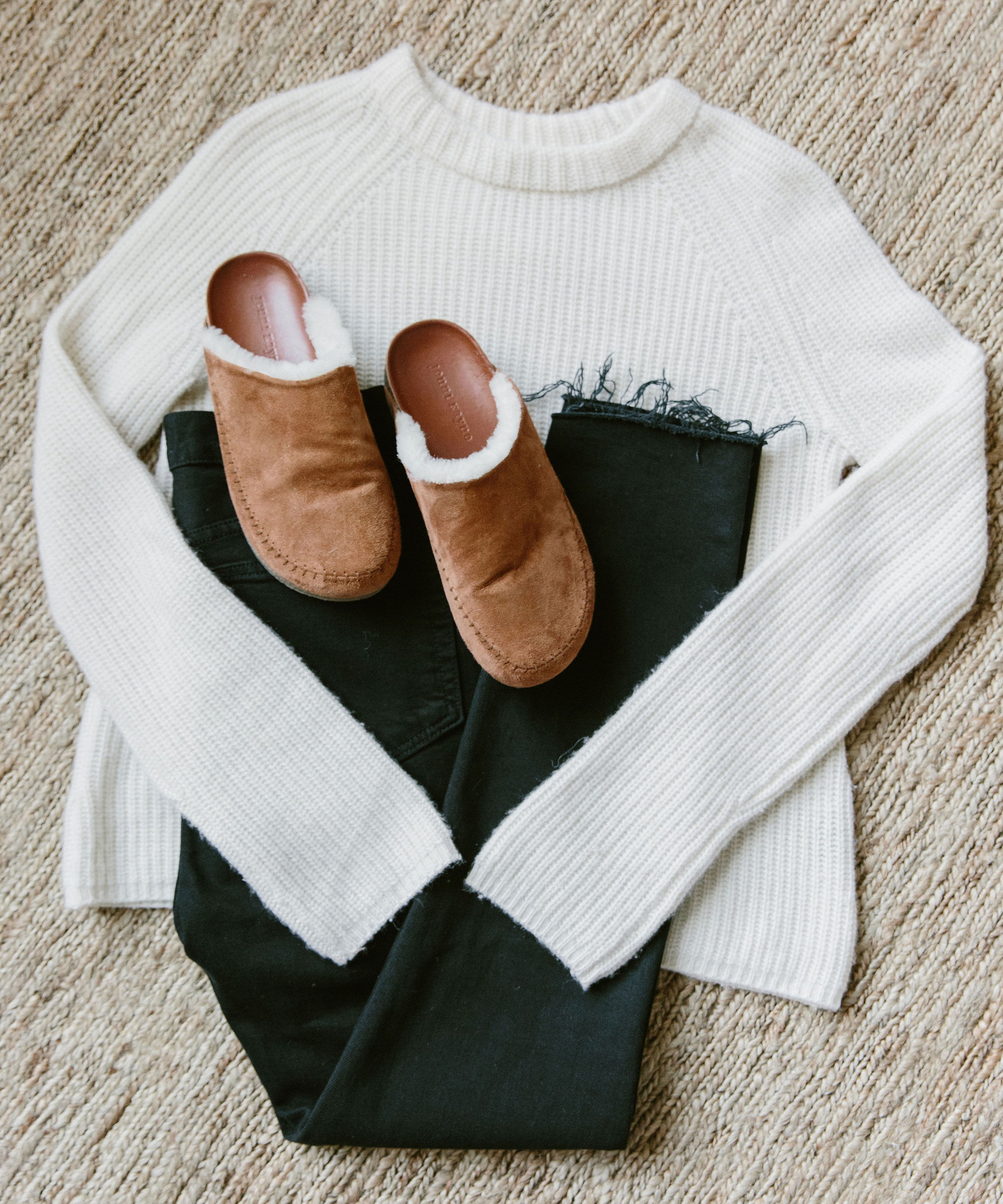 A white knit sweater, black jeans, and tan shearling-lined slippers—perfect holiday gifts—are neatly arranged on a beige textured surface.