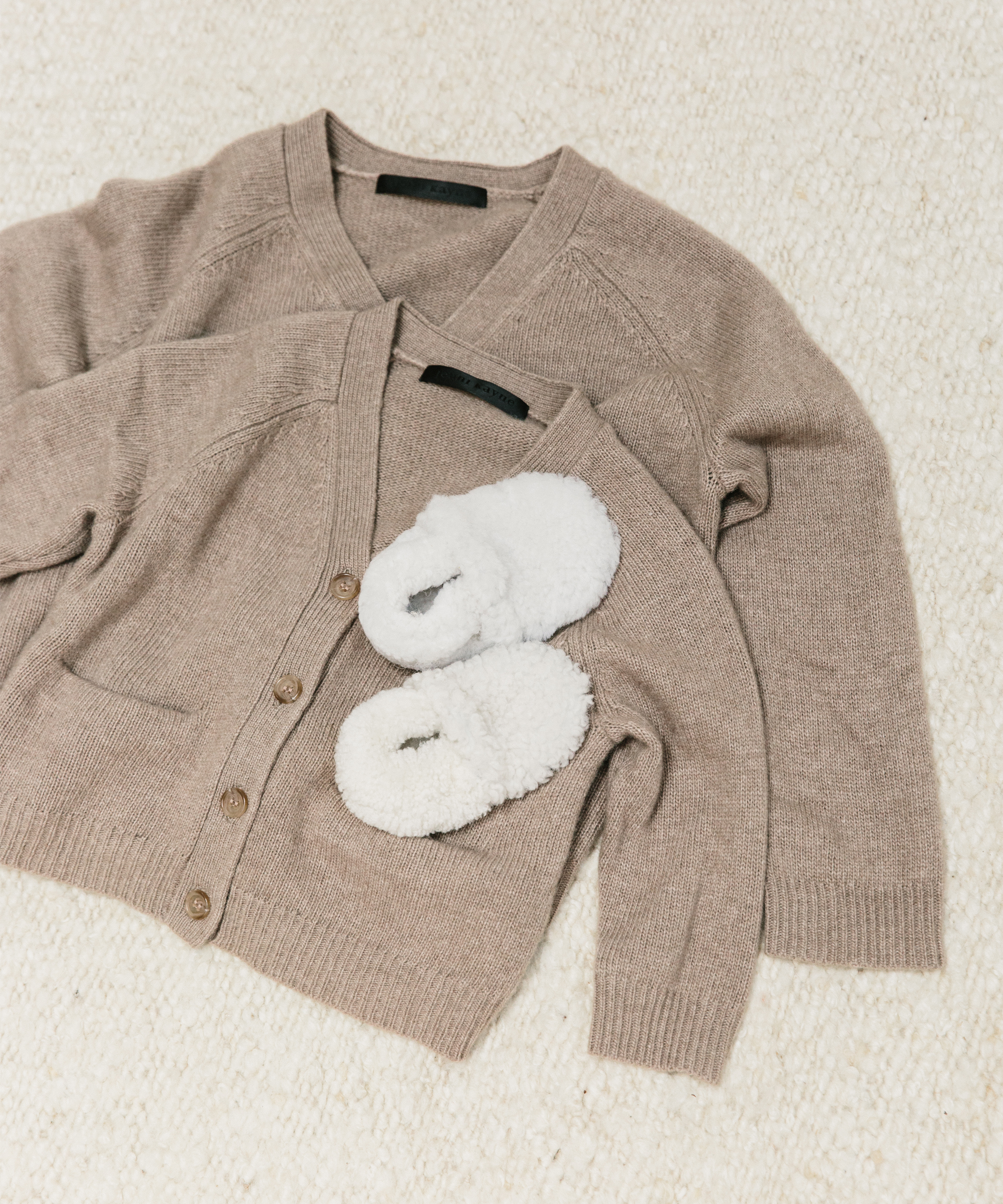 Two beige knit cardigans, one larger and one smaller, are laid out on a cream-colored textured surface. A pair of small, fluffy white slippers rests on the smaller cardigan—perfect holiday gifts for a cozy season.