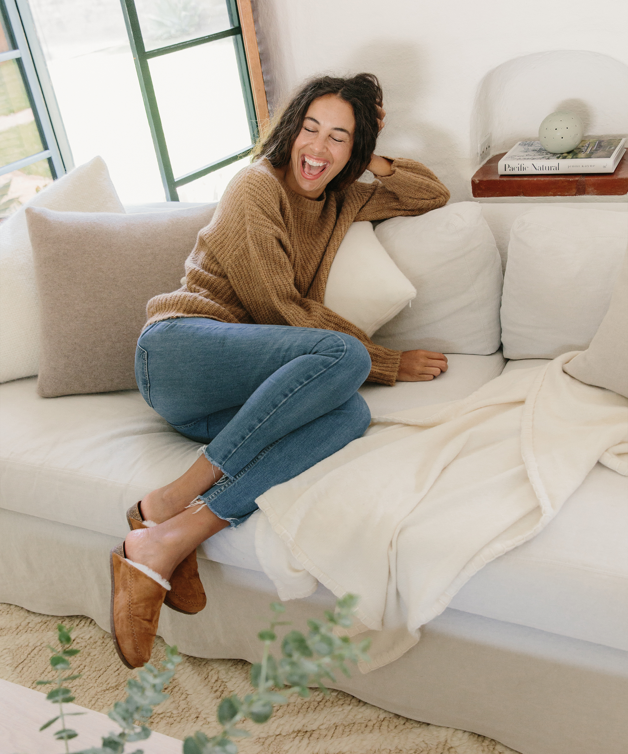 A woman in a brown sweater and jeans sits on a white couch, laughing with her eyes closed as she daydreams about holiday packing. Cozy pillows and a soft blanket add charm to the bright room, while her brown slippers complete the relaxed scene.