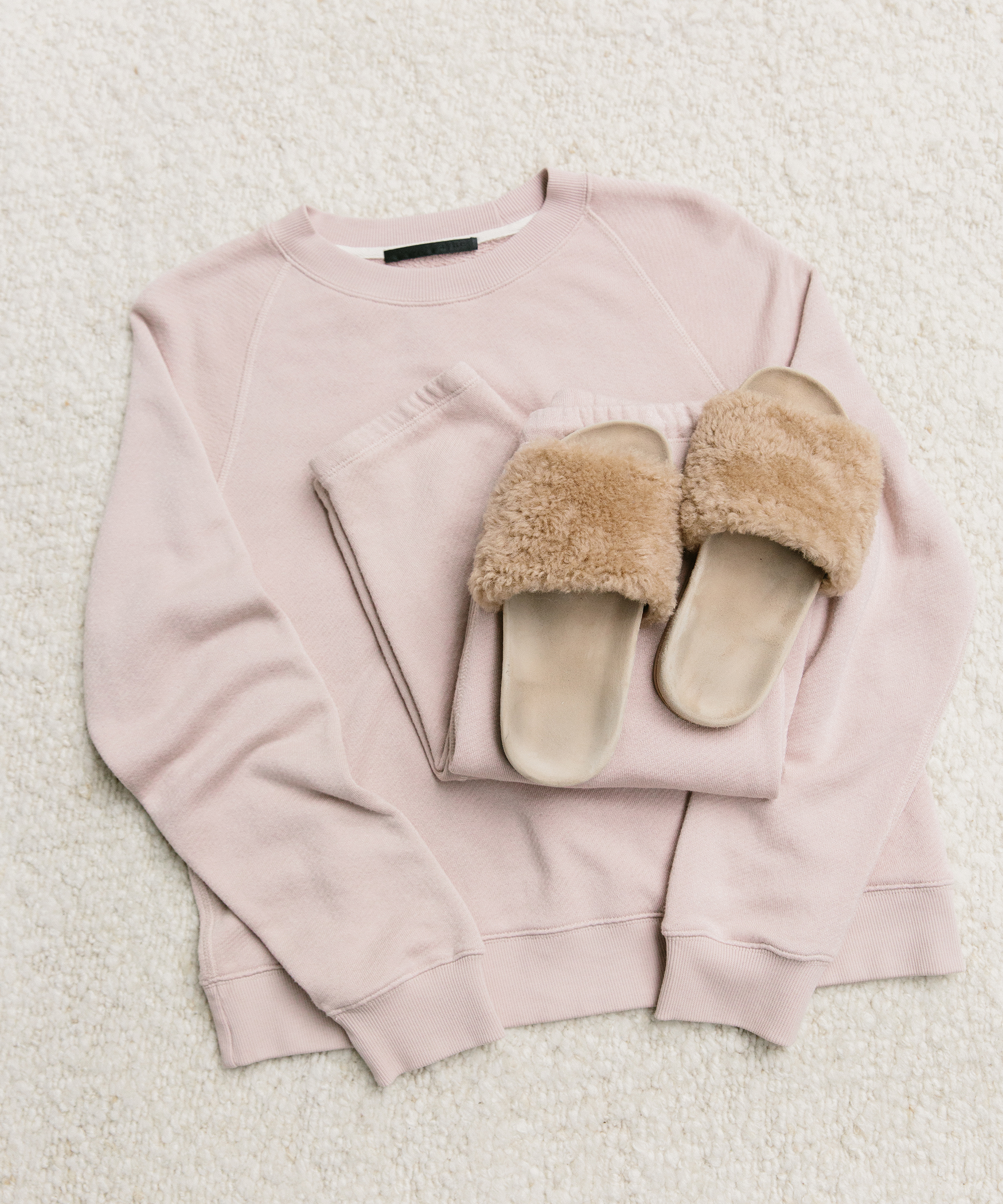 A folded light pink sweatshirt and matching sweatpants with tan fuzzy slippers placed on top, all laid out on a soft cream-colored textured surface—perfect essentials for cozy holiday packing.