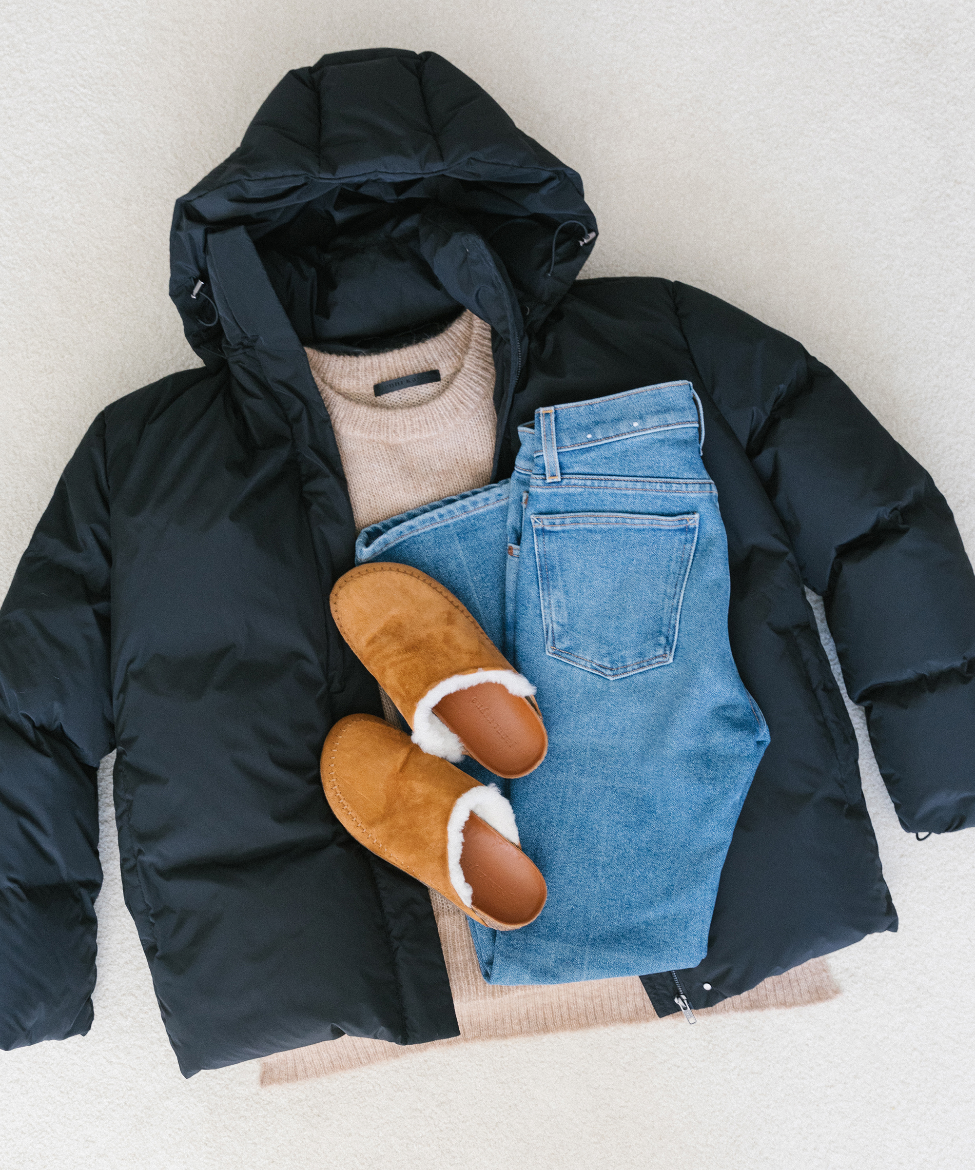 Overhead shot of a black puffer jacket with a camel sweater inside, plus saddle shearling-lined mocs, and jeans folded on top of white carpet.