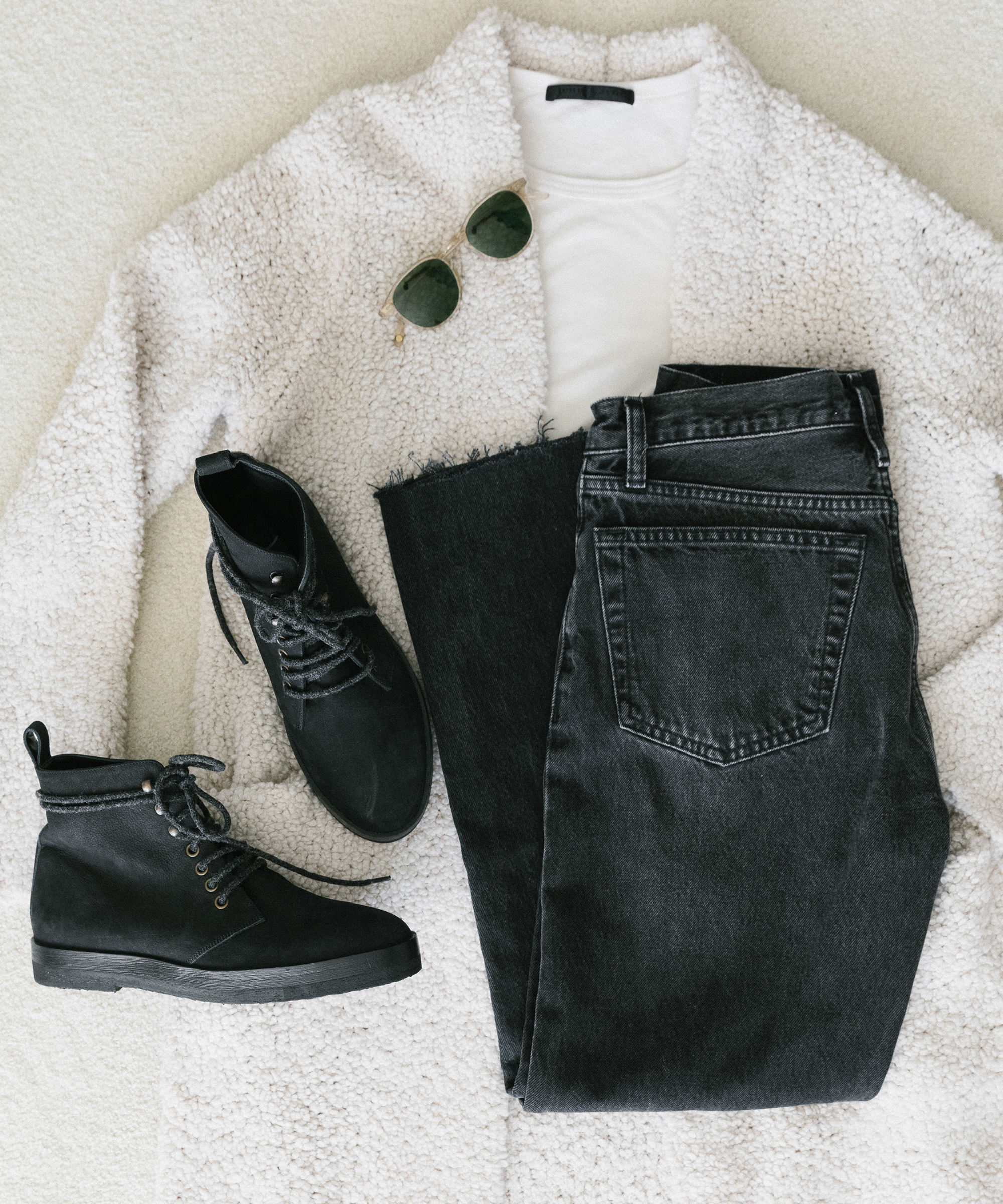 Flat lay of a cozy white sherpa coat, plain white T-shirt, black denim jeans, black lace-up ankle boots, and round green sunglasses arranged on a light surface—perfect inspiration for chic winter outfits.