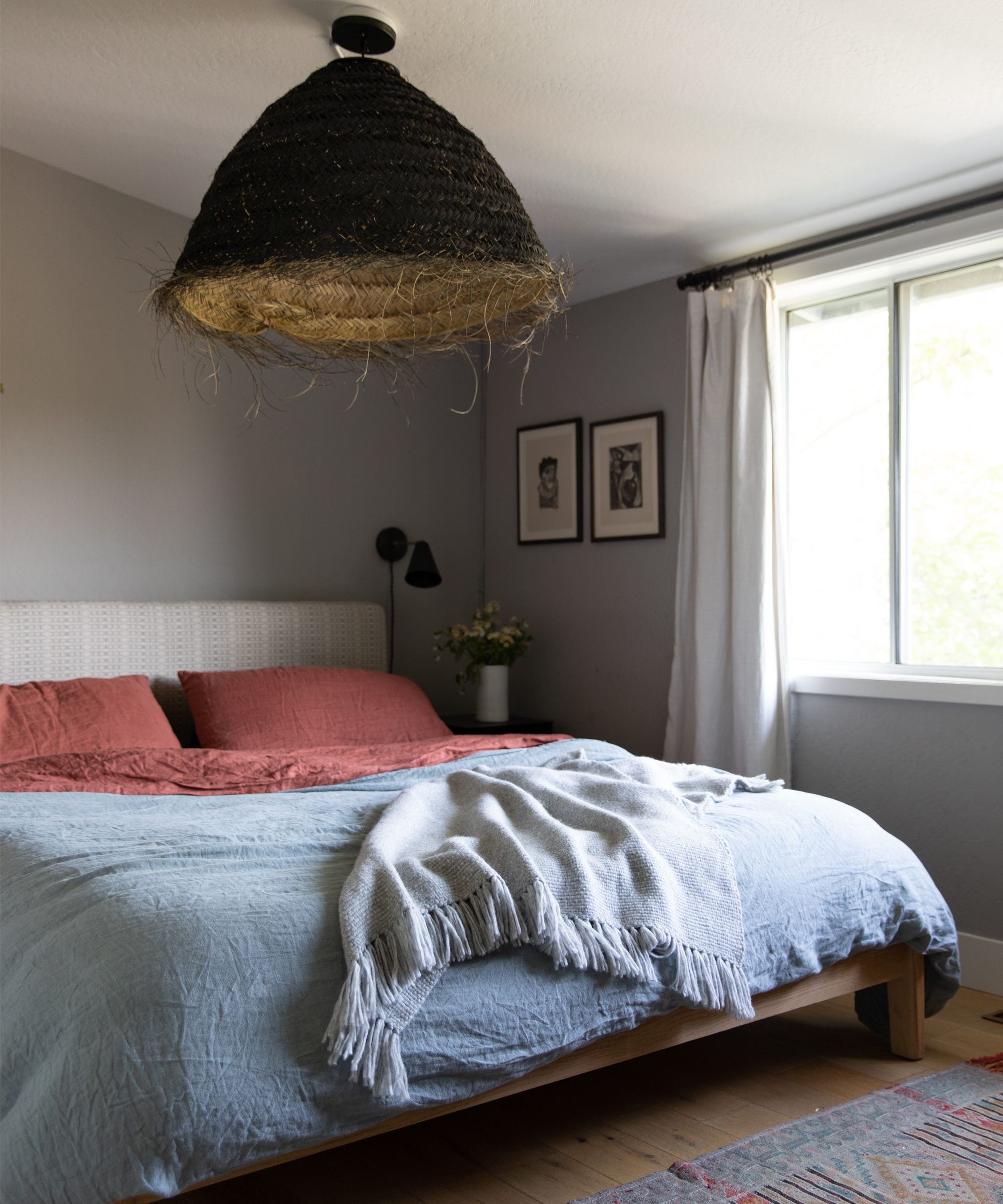 A cozy bedroom styled by Lauren Nelson Design features light blue and rust-colored bedding, a white fringed blanket, a woven pendant lamp overhead, two framed pictures on the wall, and sunlight streaming through the window.