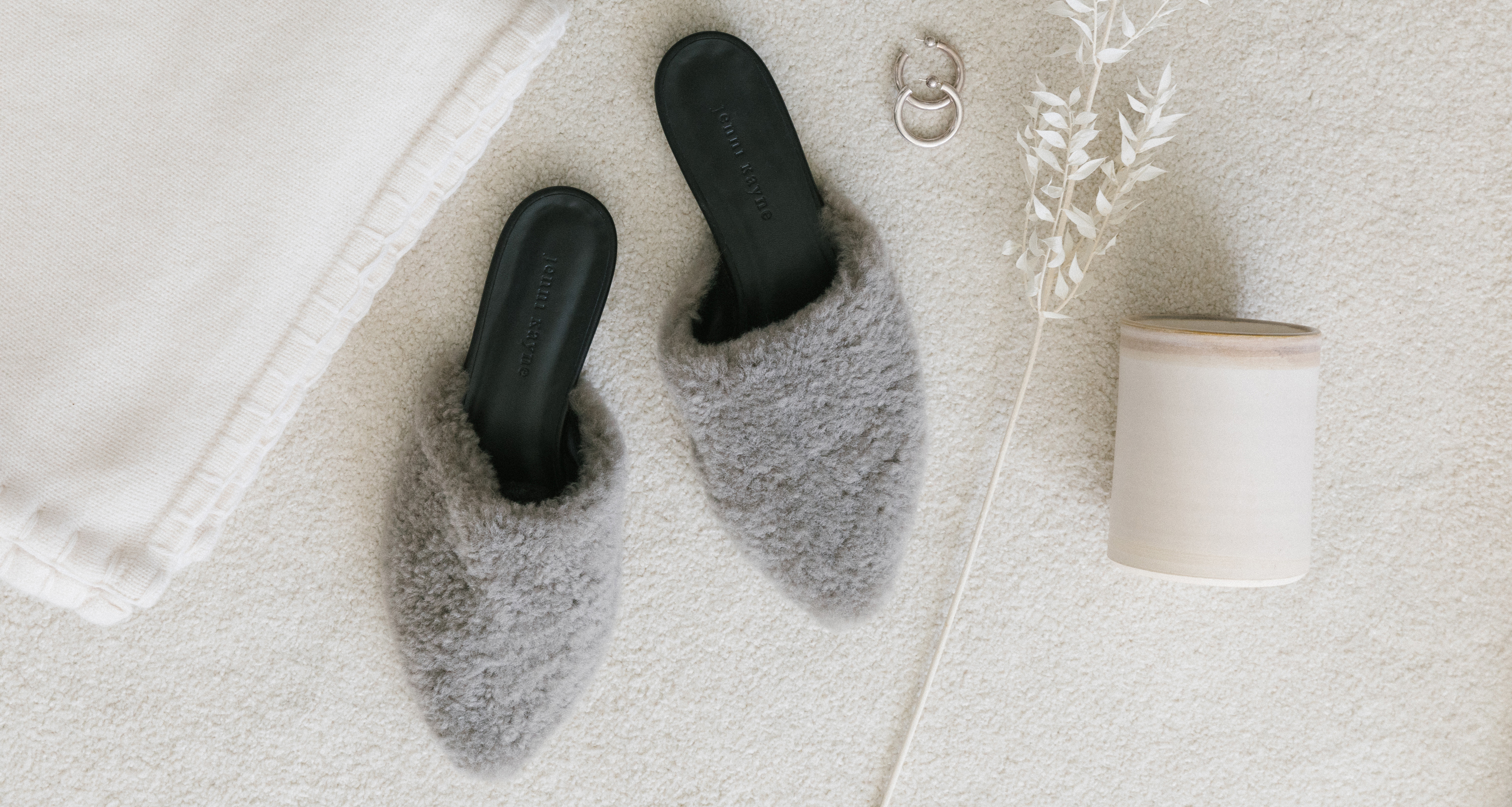 Jenni Kayne grey shearling mule, white cashmere throw, ceramic candle, earrings, and white wheat on white carpet.