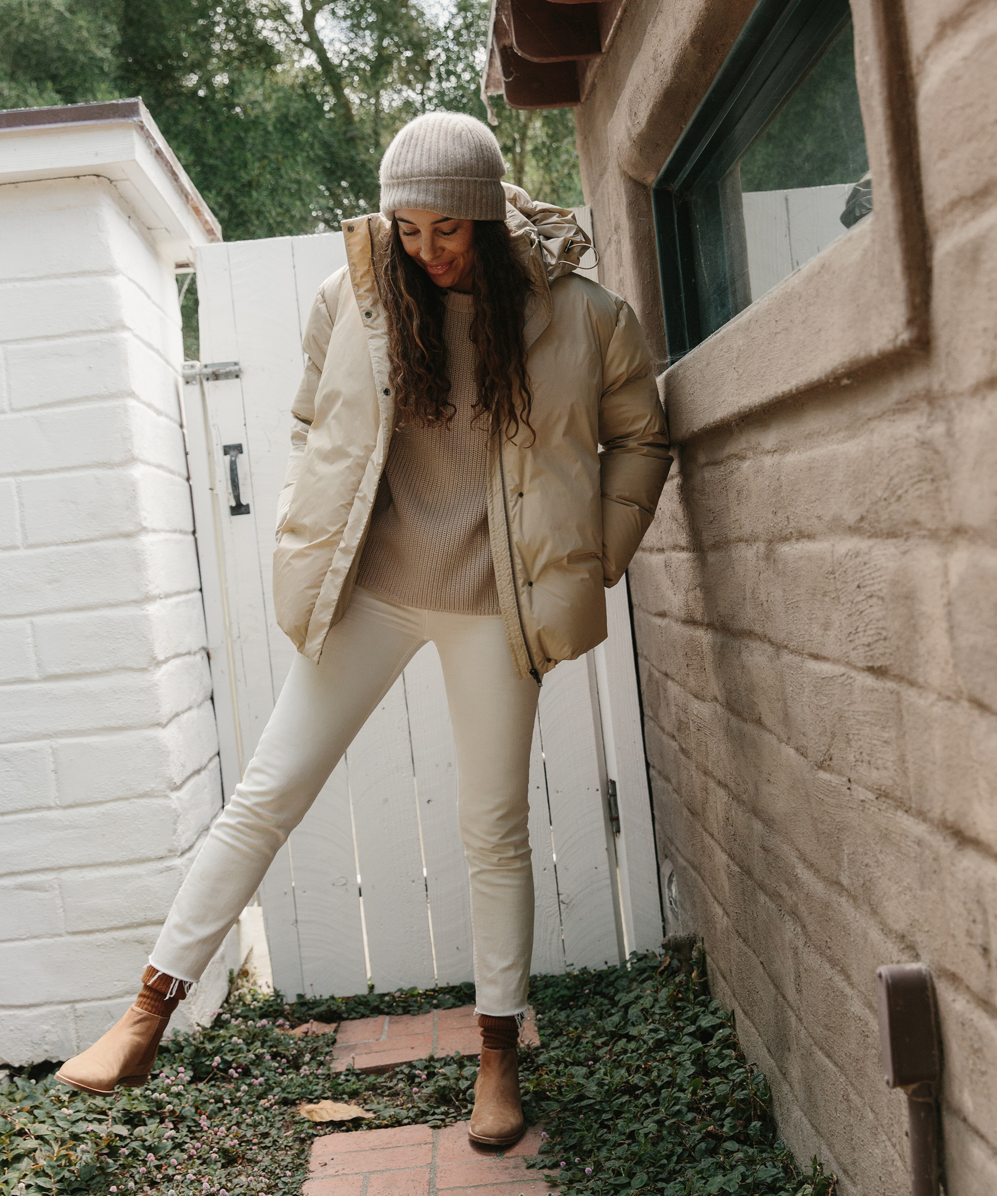 A woman wearing a beige winter coat, knit hat, sweater, white pants, and brown boots stands outdoors by a brick wall and white fence, smiling as she enjoys the crisp air—ready for her holiday packing adventures.