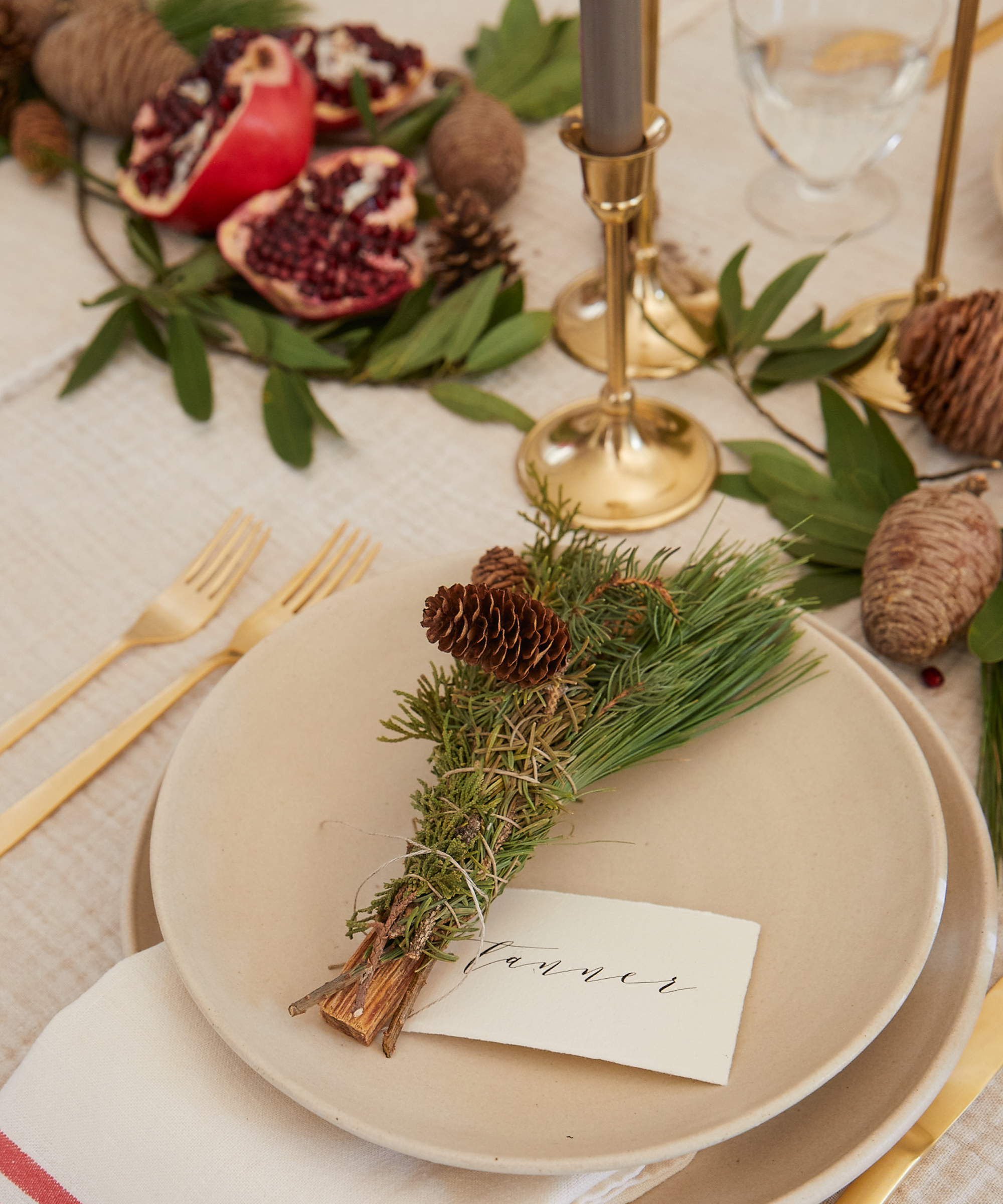 A beige plate with a small bundle of pine branches and pinecones sits beside a handwritten name card. Gold cutlery, tall gold candle holders, greenery, and split pomegranate complete this festive holiday recipes tablescape.